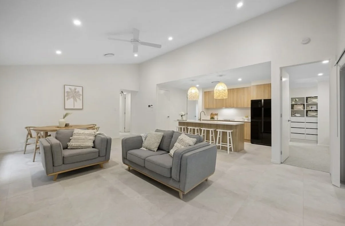 Open concept living room with gray sofas, a dining area with wooden table and chairs, and a kitchen with wooden cabinets, an island with barstools, pendant lights, and black refrigerator.