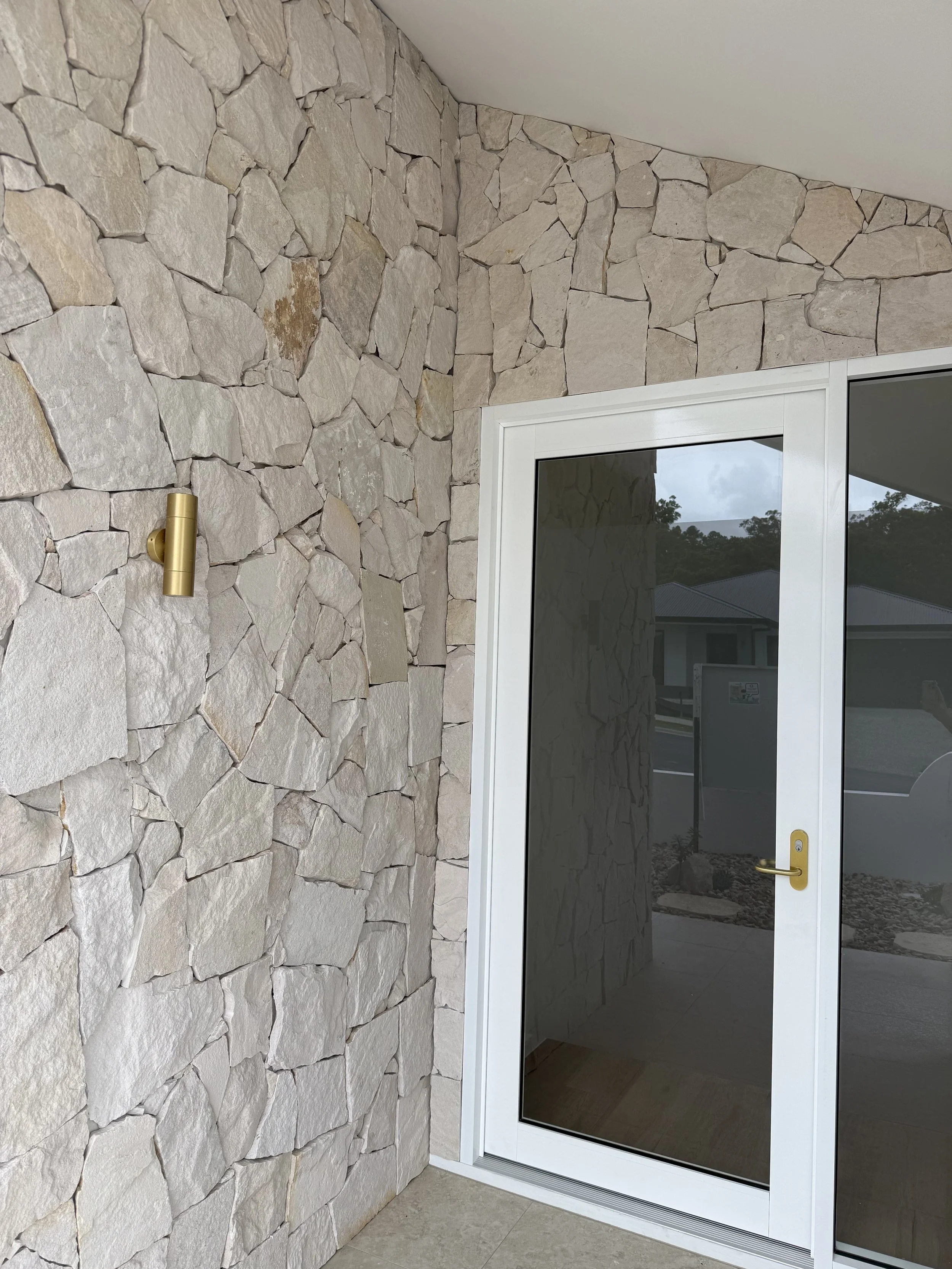 A corner of a patio or porch with a stone wall, a glass door with white frame and gold handle, and a brass outdoor wall light fixture. The stone wall has irregularly shaped beige and gray stones, and the door reflects the outdoor landscape.