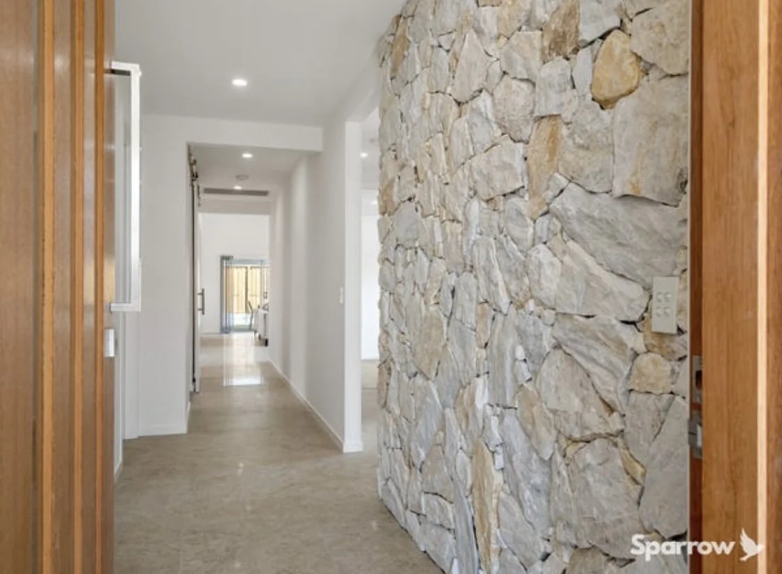 View of a hallway inside a modern home with a stone accent wall on the right and white walls, with a door opening to a room with sliding glass doors at the far end.