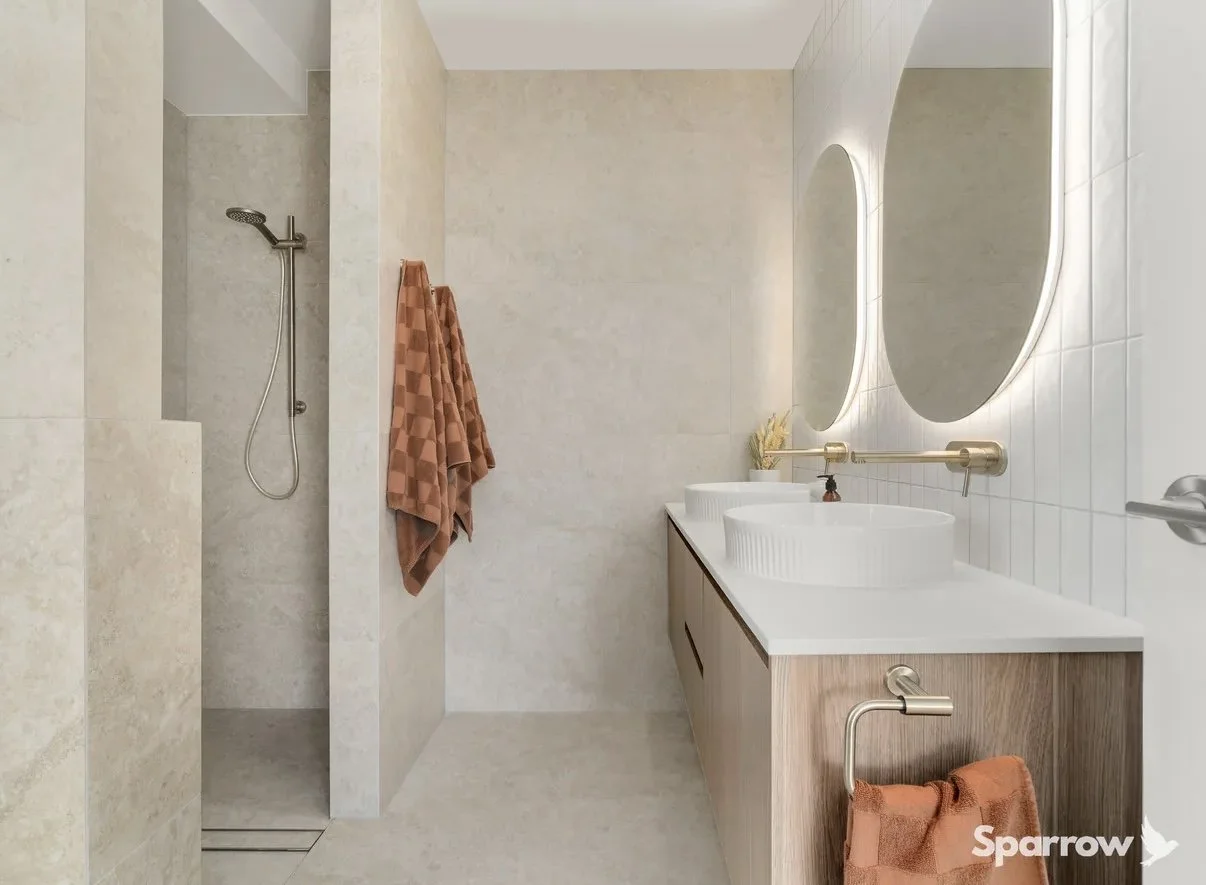 Modern bathroom with walk-in shower, two round mirrors above double sinks, wooden vanity, rust-colored towels, and minimal decor.