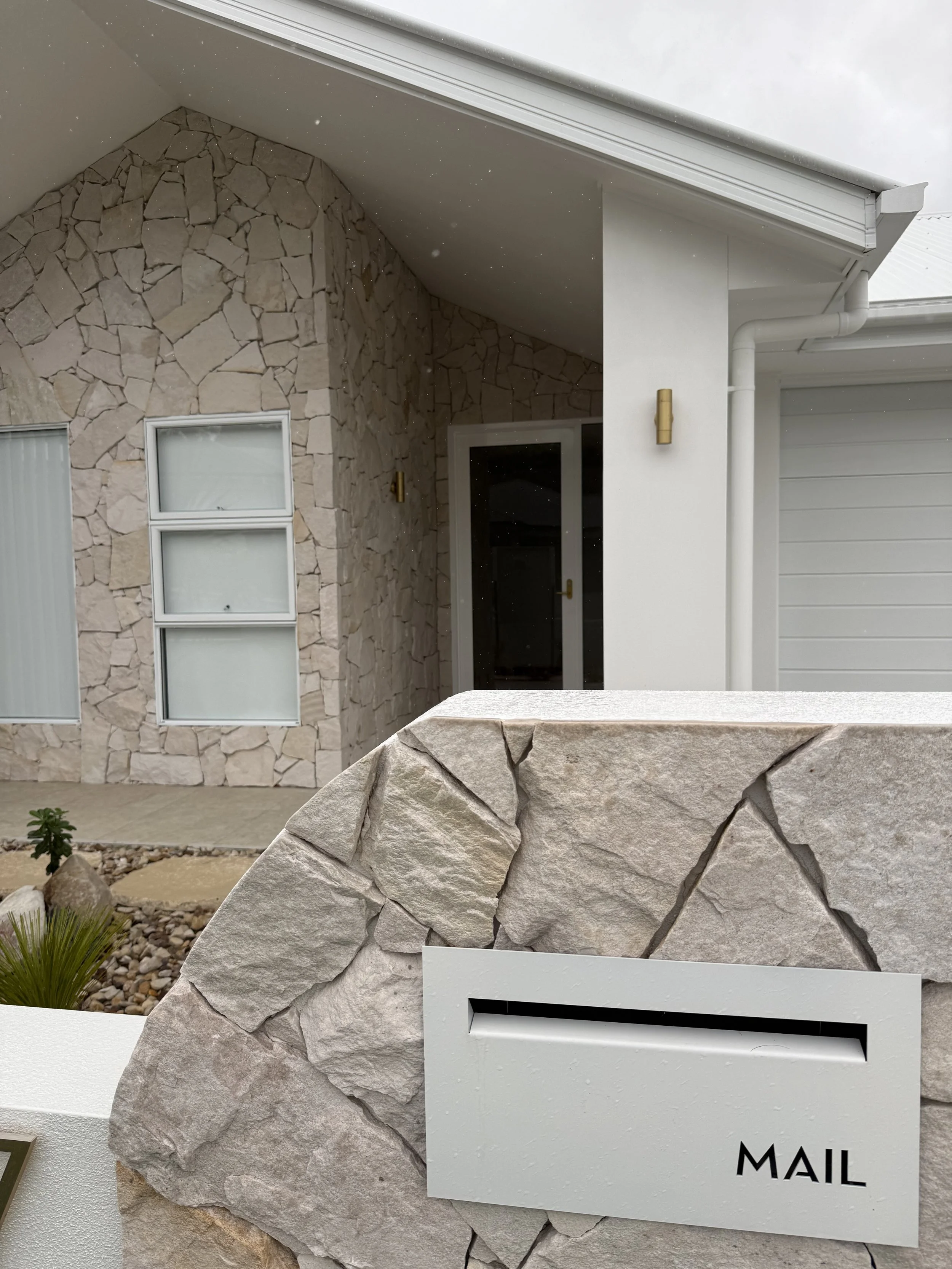 Close-up of a modern house exterior with stone and white wall, glass window, gold light fixtures, and a white mailbox labeled 'MAIL'
