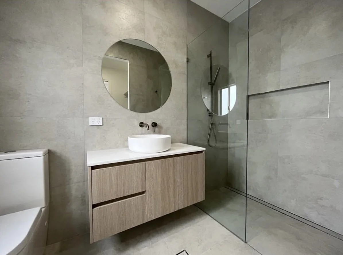 Modern bathroom with a floating wooden vanity, a round mirror, a vessel sink, a wall-mounted faucet, a toilet, and a glass shower with a wall niche and a rain showerhead.