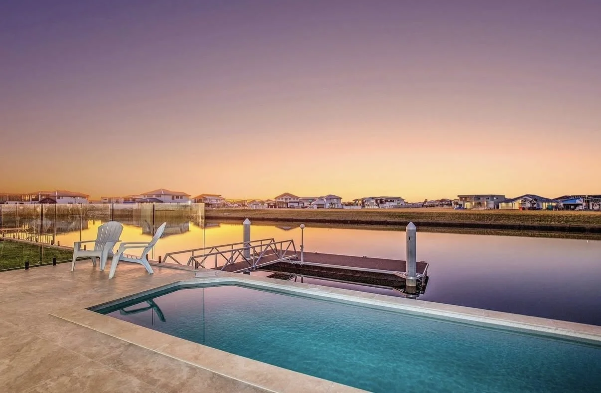 Sunset view of a backyard pool overlooking a calm waterway with houses in the distance, two white chairs on the patio.