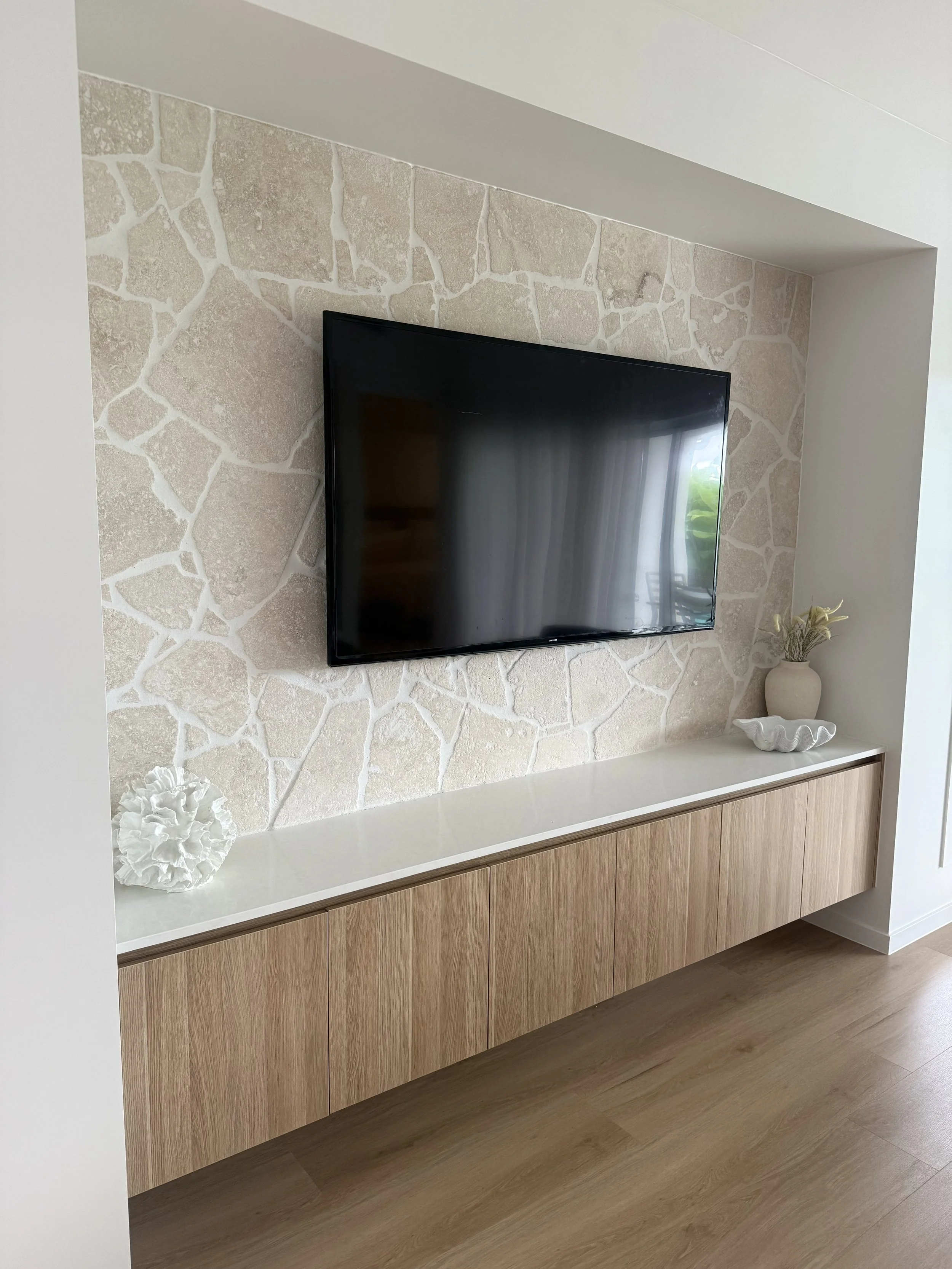 Living room wall with textured stone accent, mounted flat-screen TV, a white shelf with decorative objects including a vase with dried plants and a white sculptural piece, and wood flooring.
