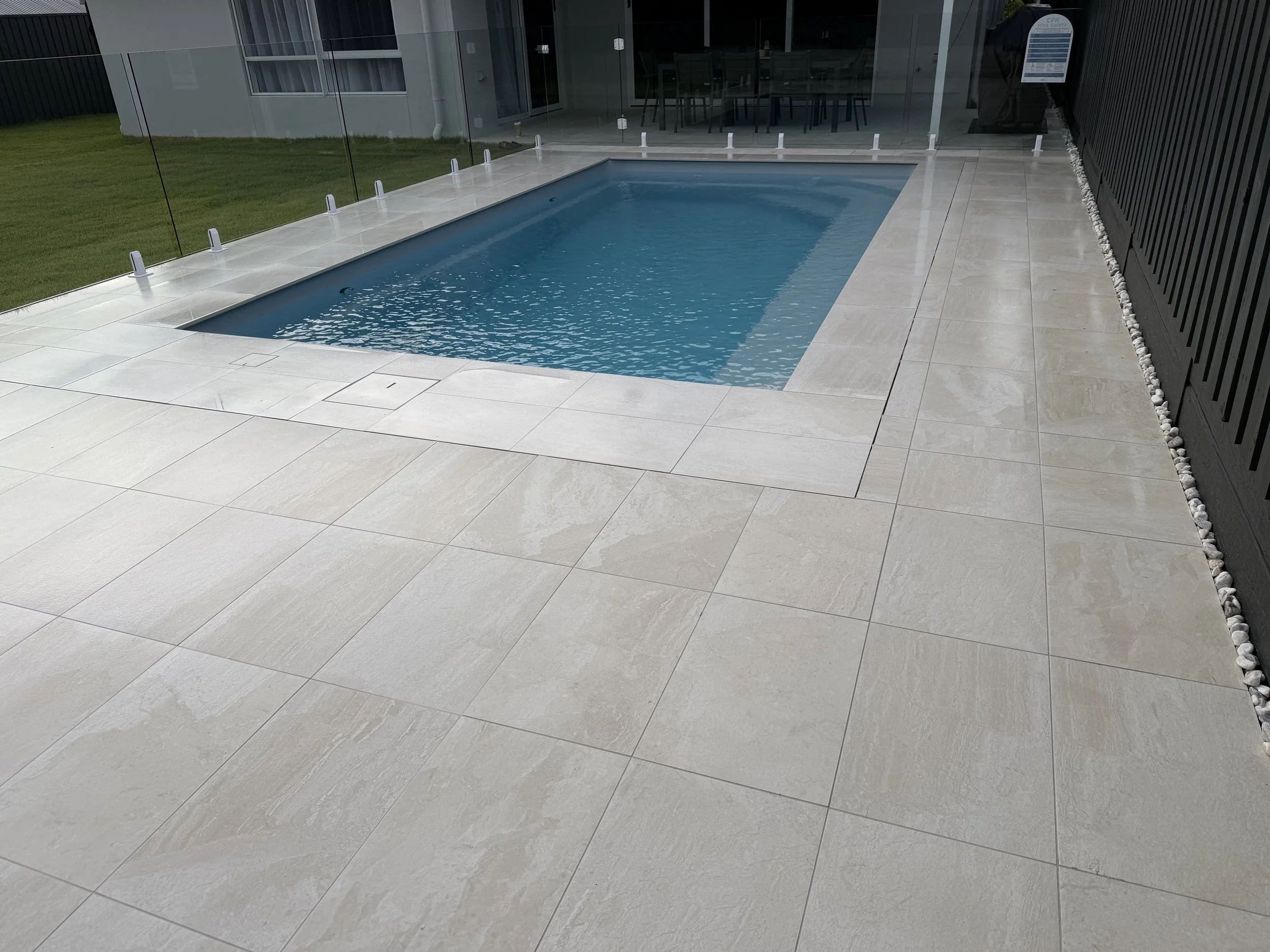 Empty rectangular swimming pool with light gray tile deck, enclosed by glass fencing, next to a house with a patio and dining area visible through the glass.