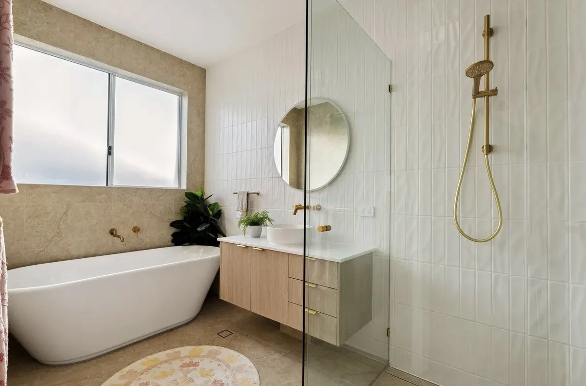 Bathroom with a bathtub under a large window, a floating vanity with a vessel sink and round mirror, a plant on the vanity, and a separate shower area with a glass partition and gold fixtures.