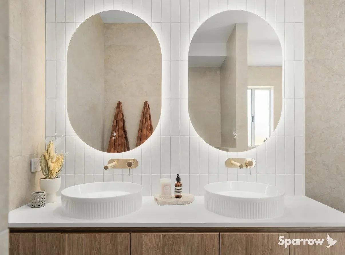 Bathroom vanity with two round vessel sinks, large oval mirrors, a small vase with dried flowers, and a bottle of soap on a tray. Wooden cabinet below. Beige tiled walls.