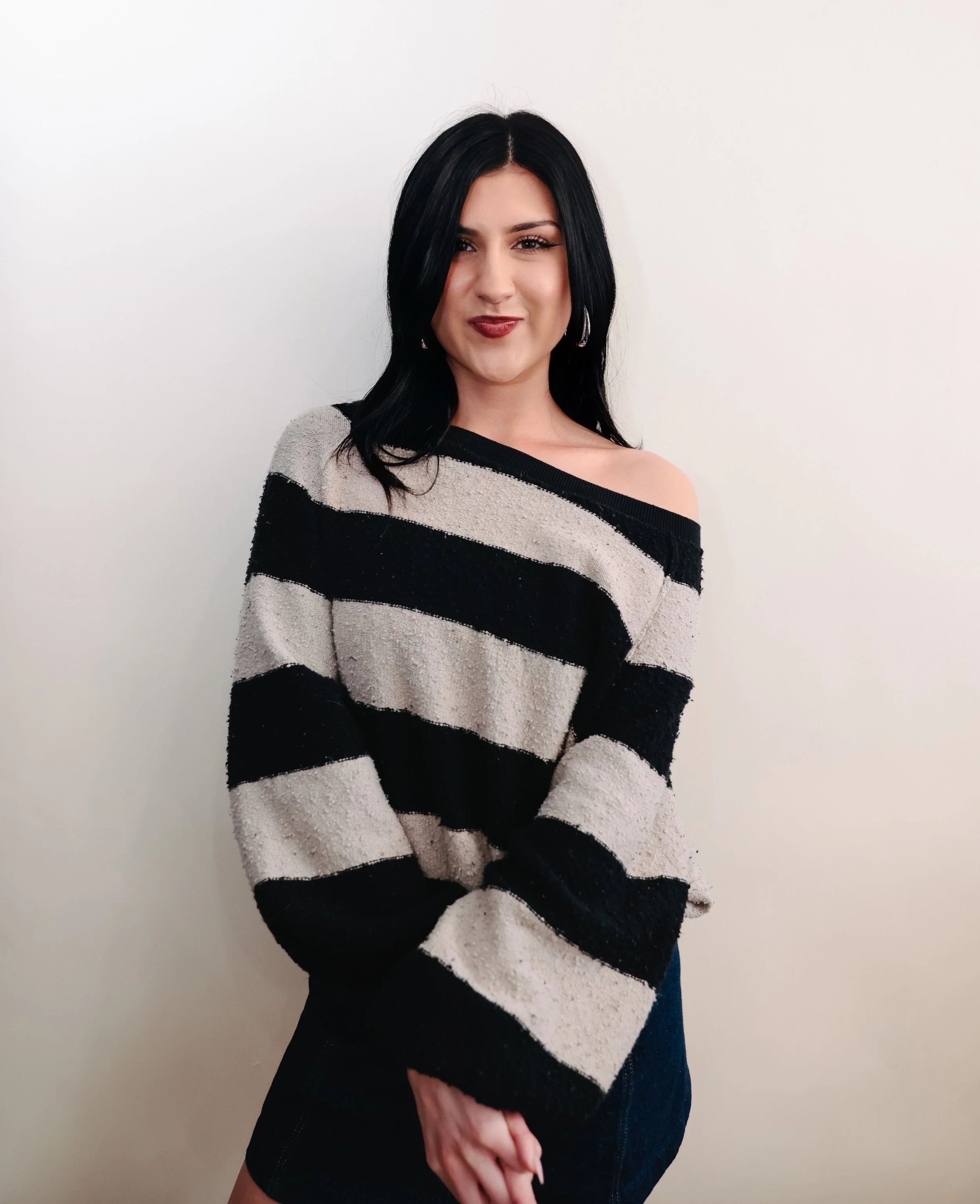 A young woman with long black hair and a subtle smile, wearing a black blouse and blue jeans, standing against a plain white background.