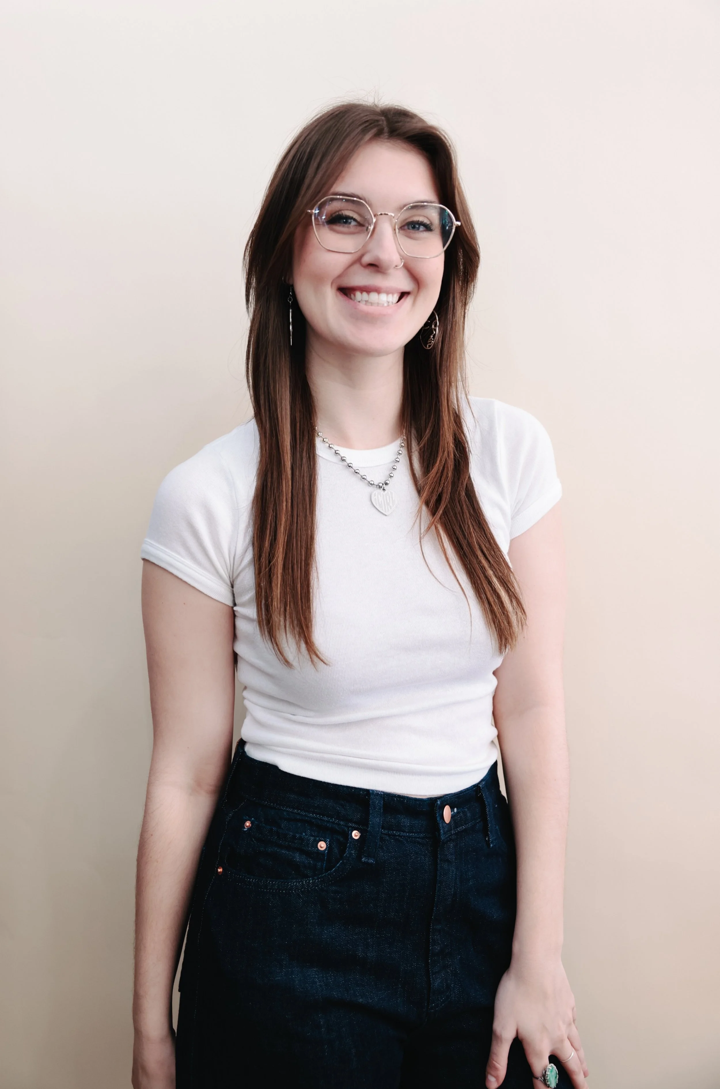 A young woman with long brown hair, wearing glasses, a cream-colored ribbed off-the-shoulder sweater, and light blue high-waisted jeans, standing against a plain light background.
