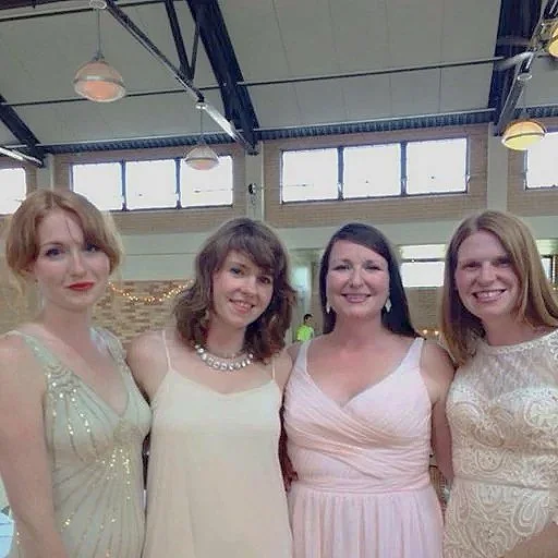 Four women in formal dresses pose together indoors in a well-lit space with high windows and ceiling lights.