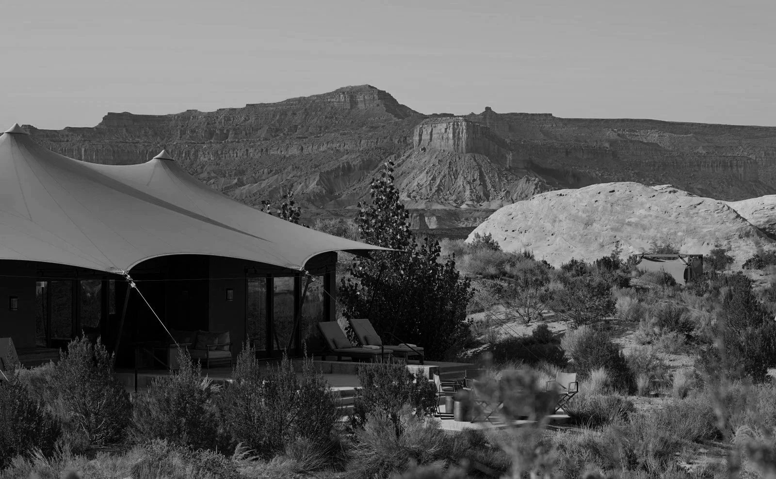 Black and white photo of a tent-like structure with outdoor seating, set against a rocky desert landscape with mesas and sparse vegetation.