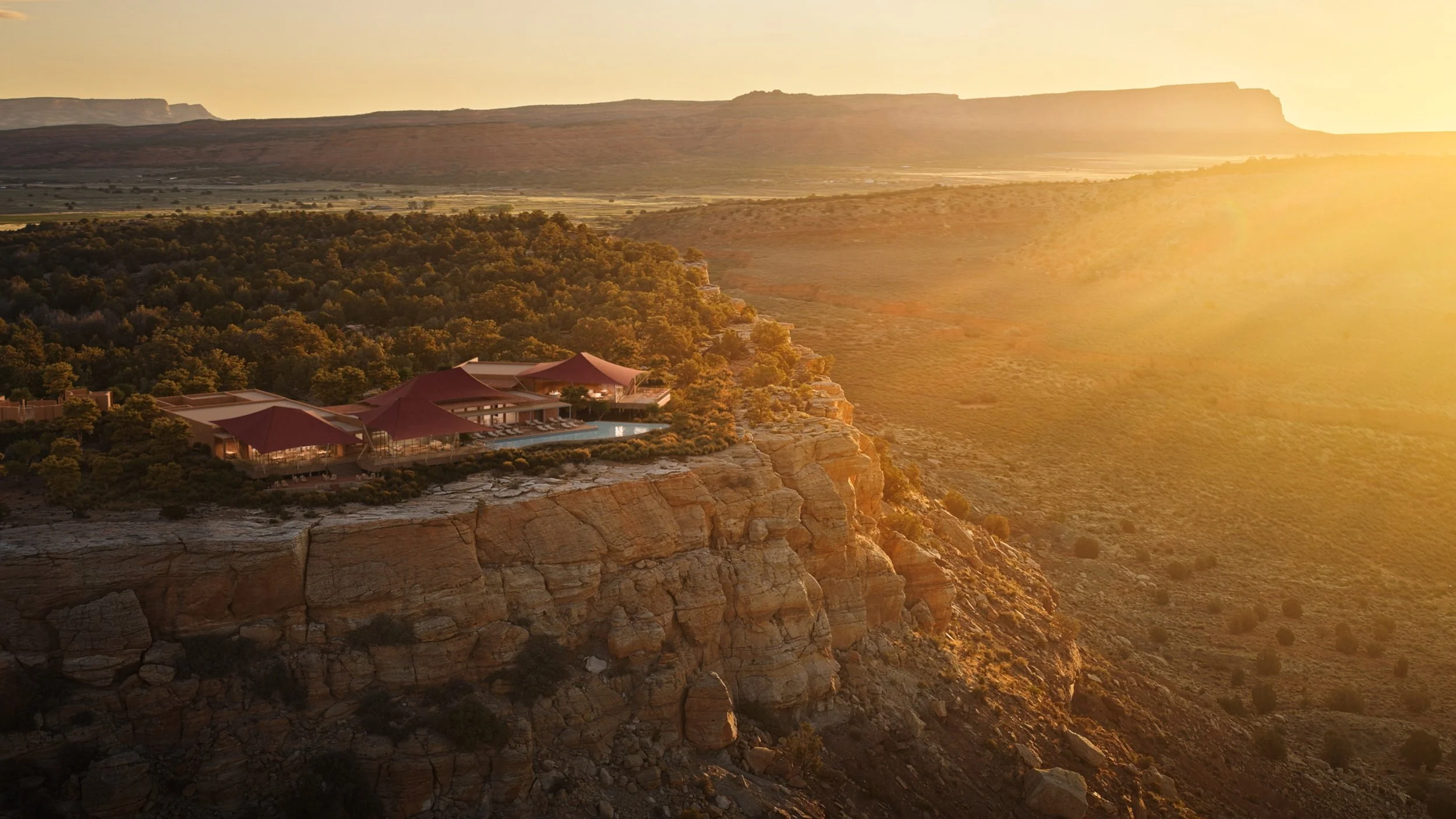 A large building with a red roof and outdoor swimming pool located on the edge of a rocky cliff overlooking a vast, flat landscape at sunset.