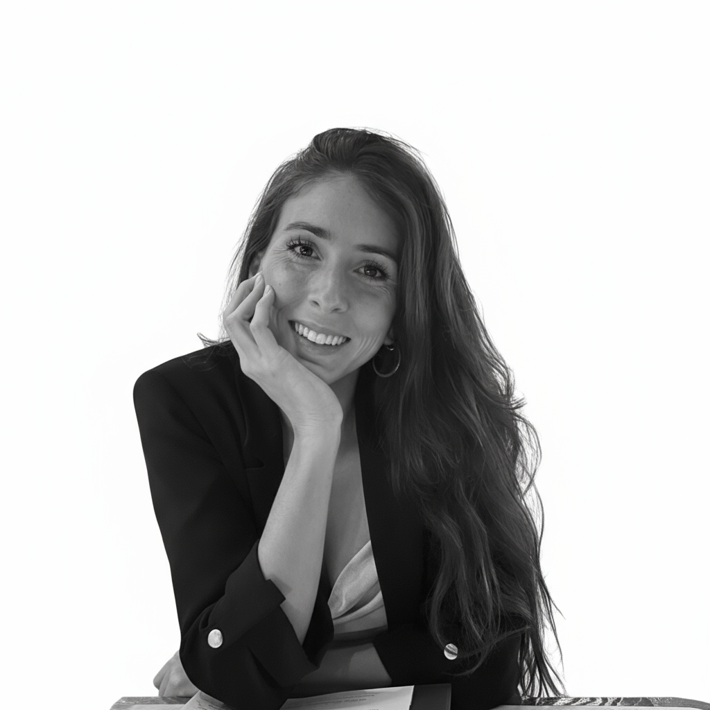 A woman with long, wavy hair smiling, resting her chin on her hand, sitting at a desk, wearing a dark blazer and a light shirt, in a black and white photo.