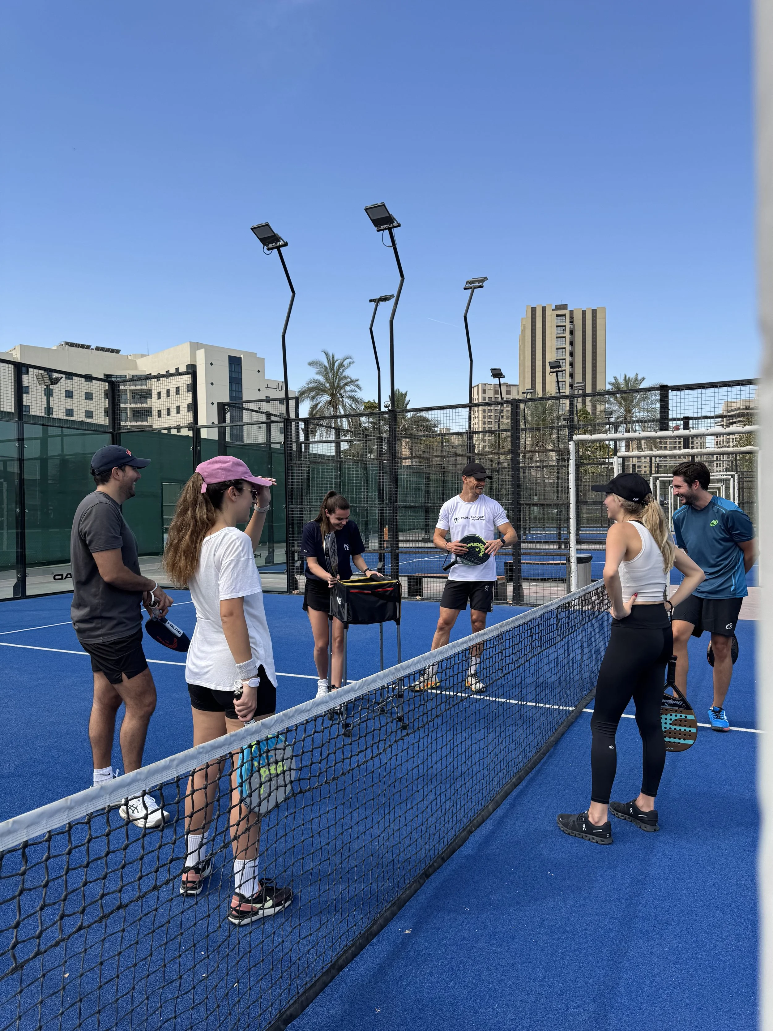 Group of people on a blue padel tennis court, standing around the net, with palm trees and tall buildings in the background, under a clear sky.