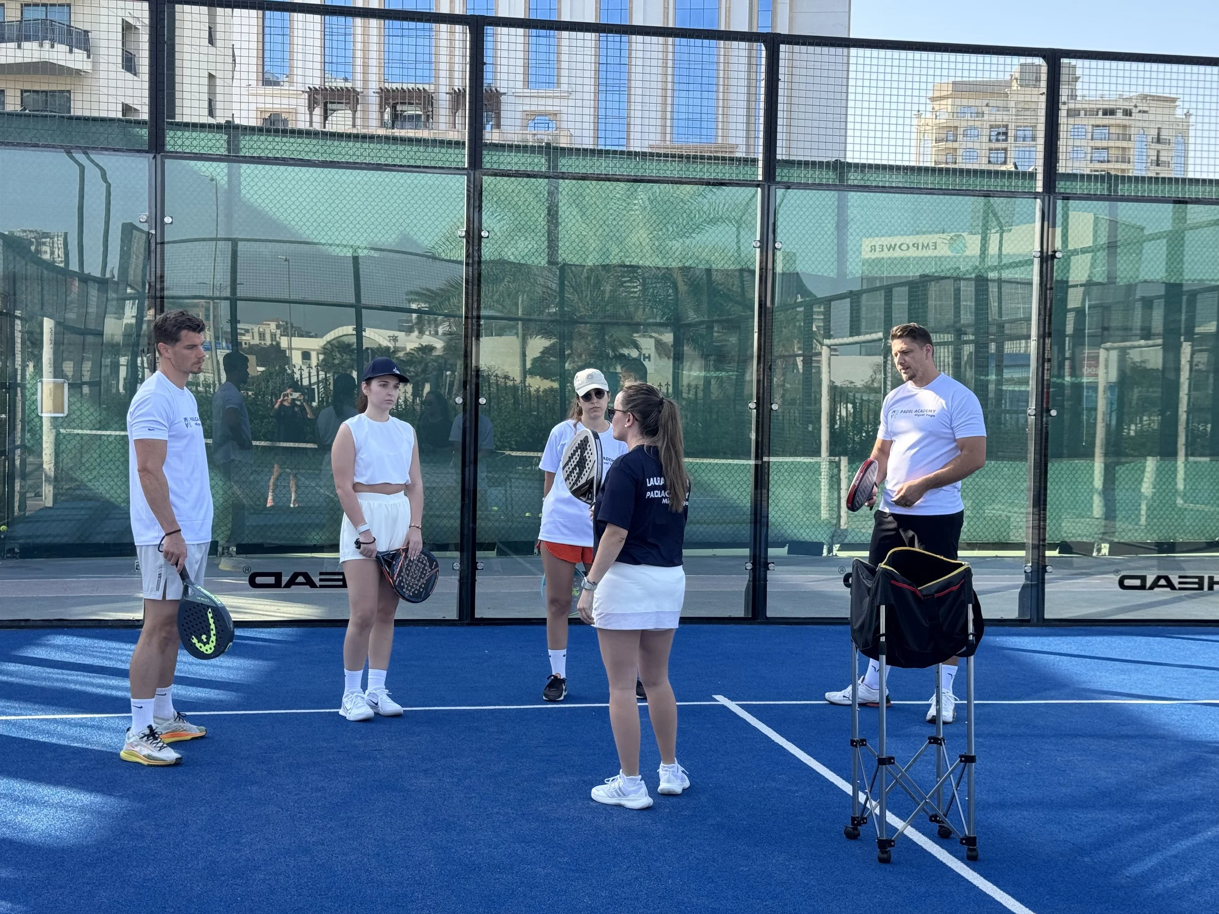 Group of people on a blue paddle tennis court with a coach giving instructions.