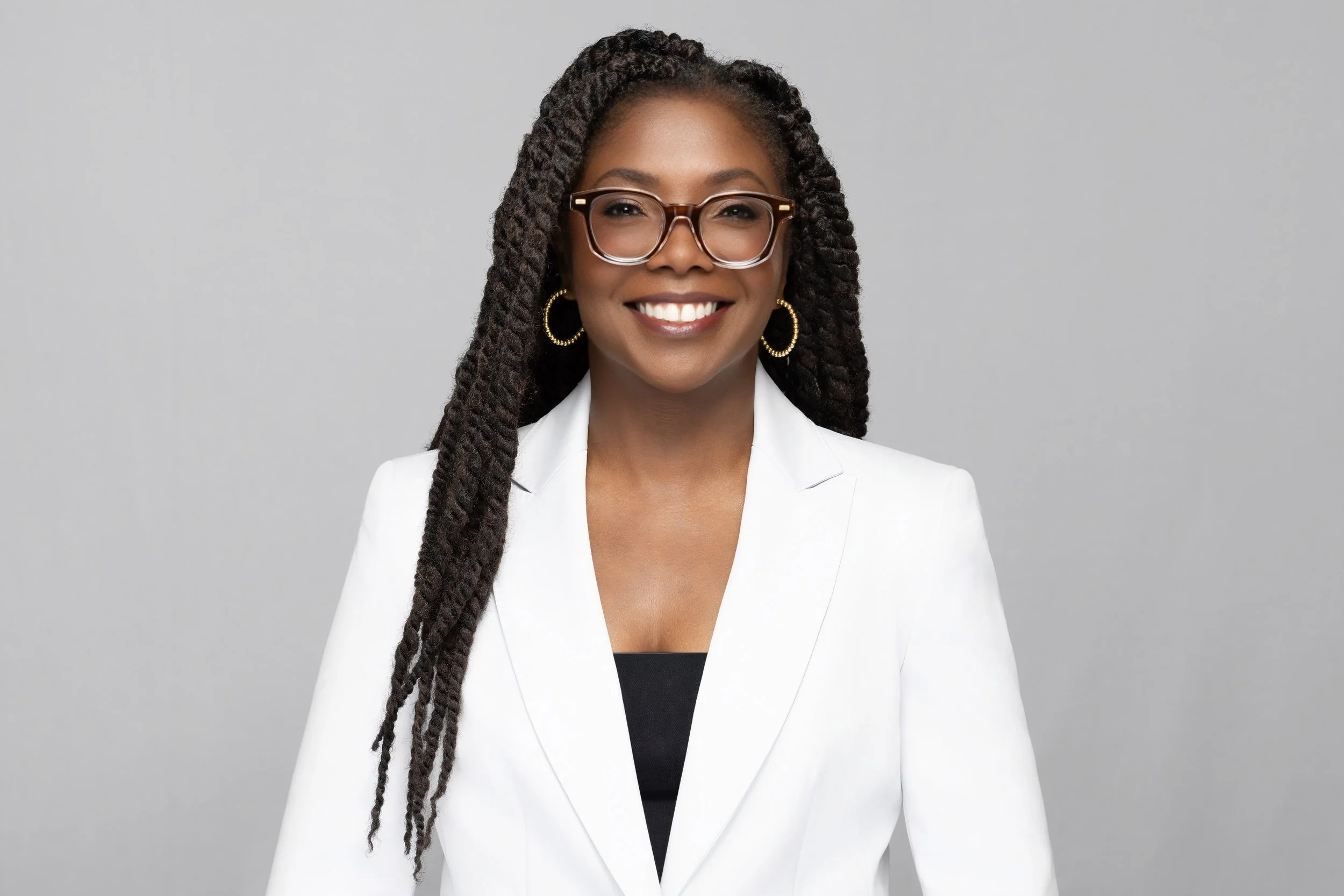 A woman with dark skin and long braided hair wearing glasses, gold hoop earrings, a white blazer, and a black top, smiling against a plain gray background.