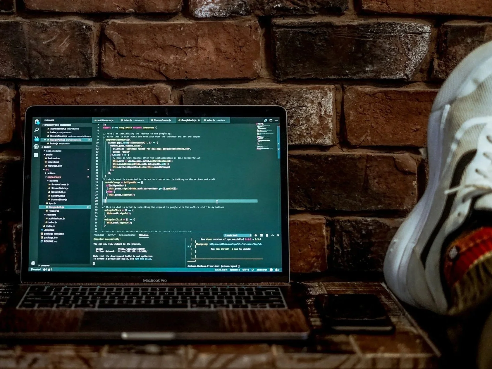 A MacBook Pro laptop on a wooden table with coding on the screen, next to a smartphone, against a brick wall background.