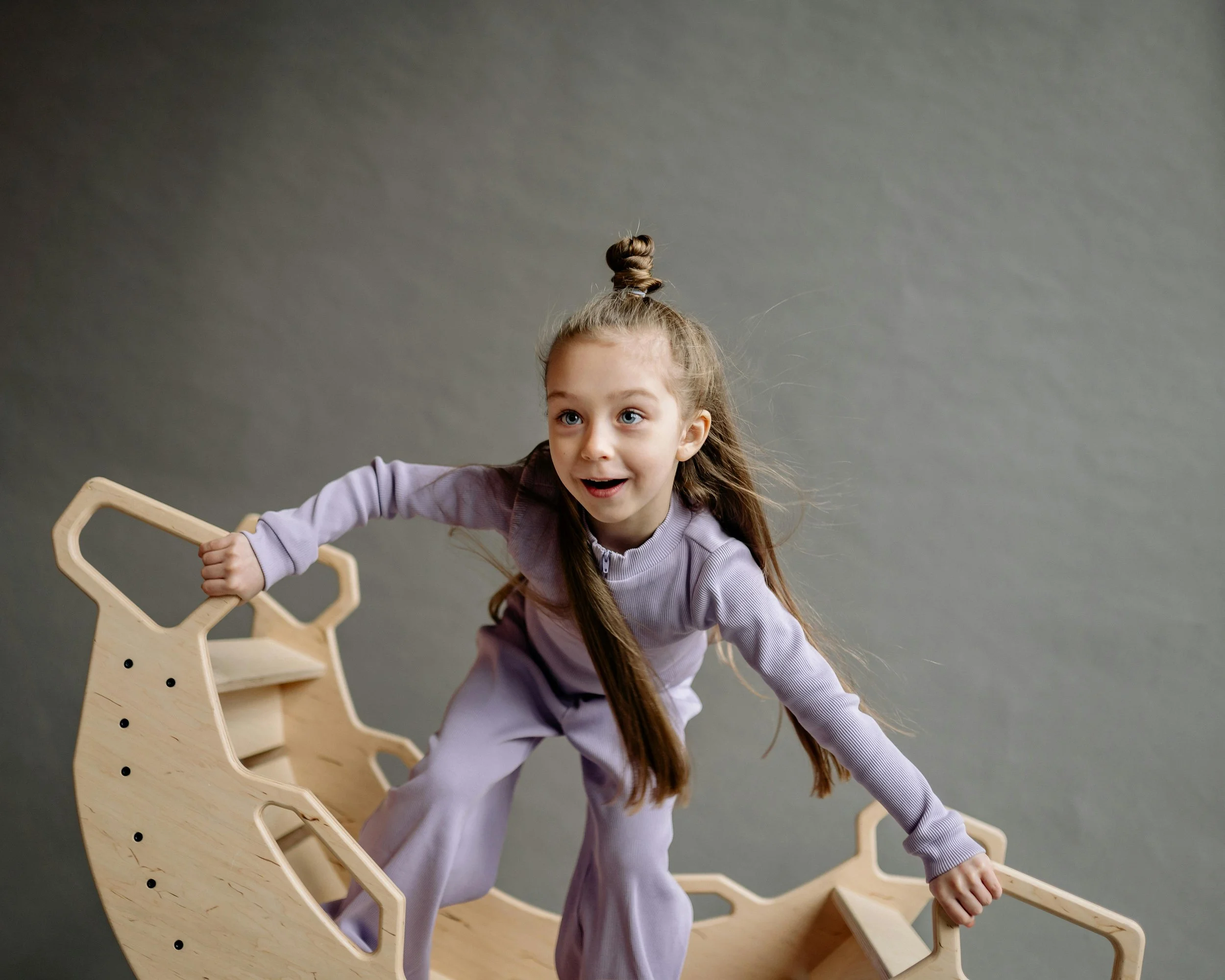 Young girl with a top knot hairstyle wearing a purple tracksuit, smiling and climbing on a wooden crescent-shaped rocking toy.