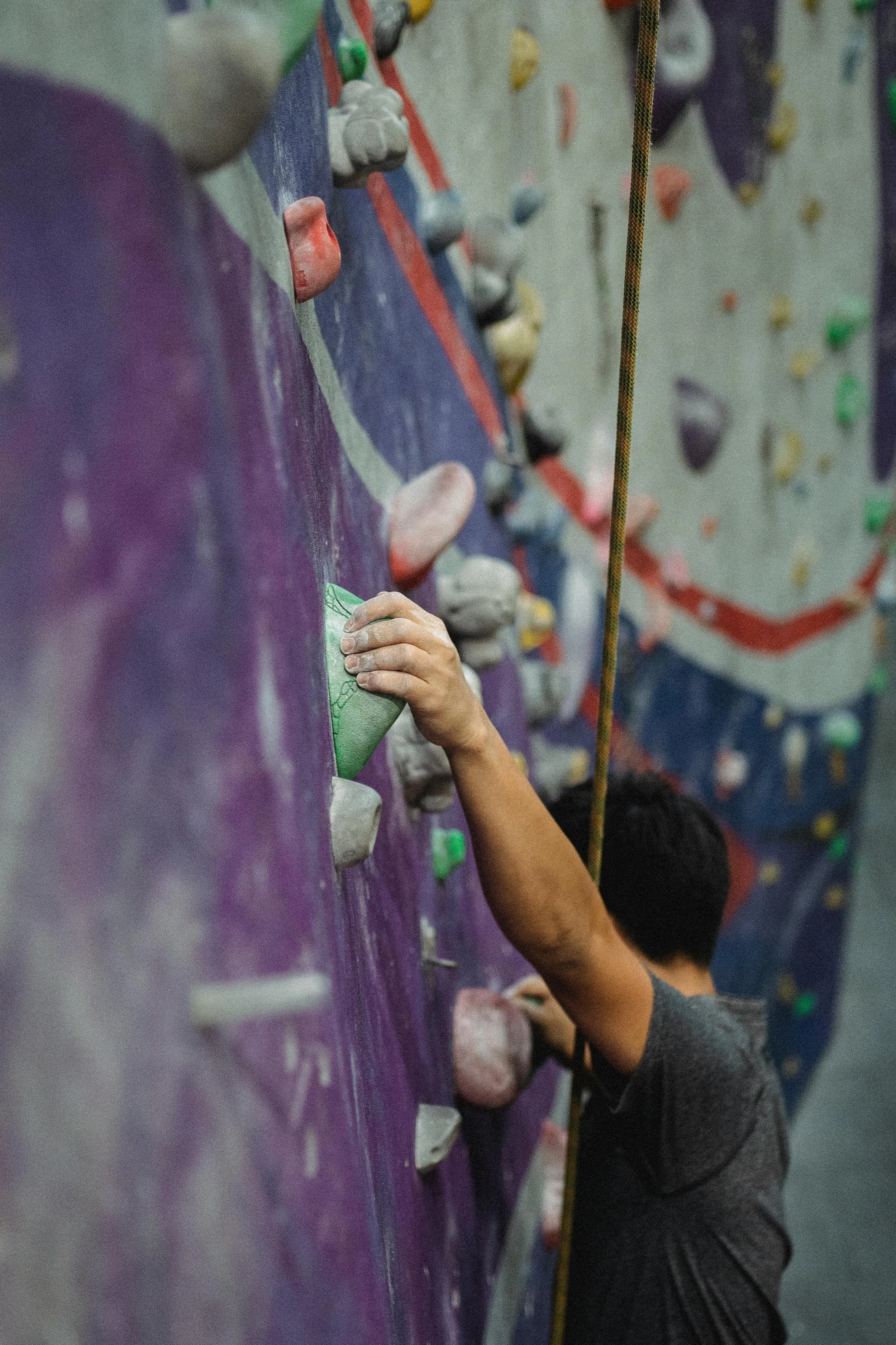 Person climbing an indoor rock climbing wall, gripping a green hold with their right hand.