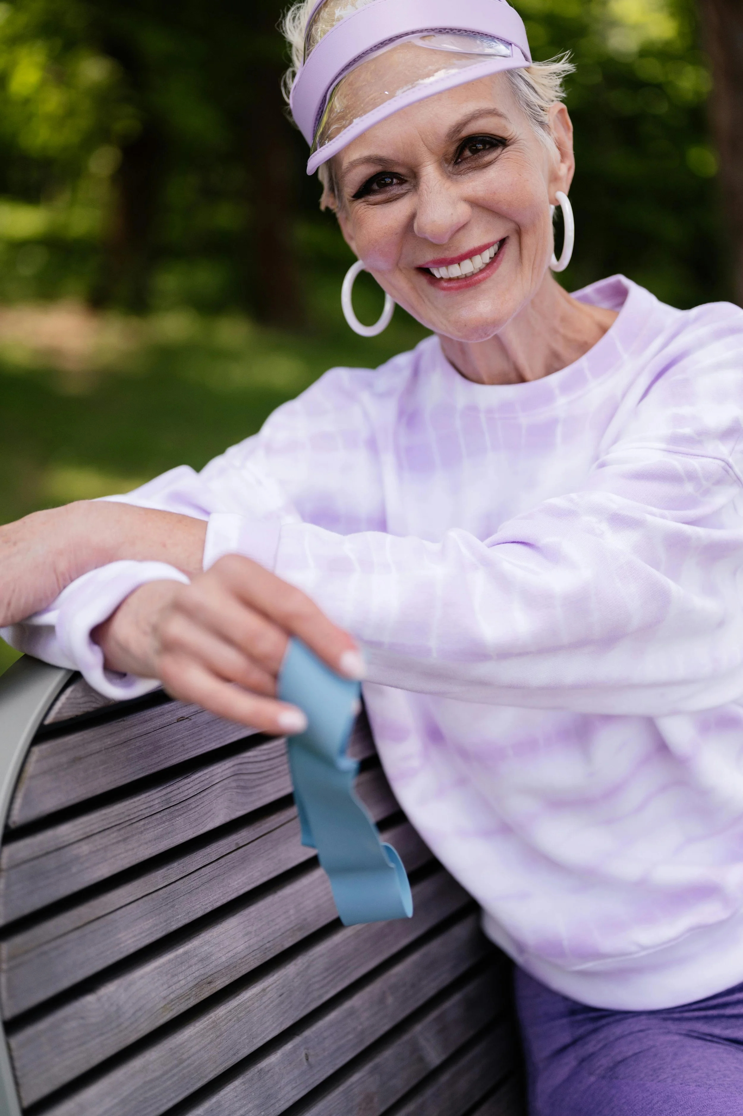 An elderly woman with short white hair wearing a lavender visor, white hoop earrings, and a light purple checkered shirt sitting on a park bench, smiling, holding a blue resistance band.