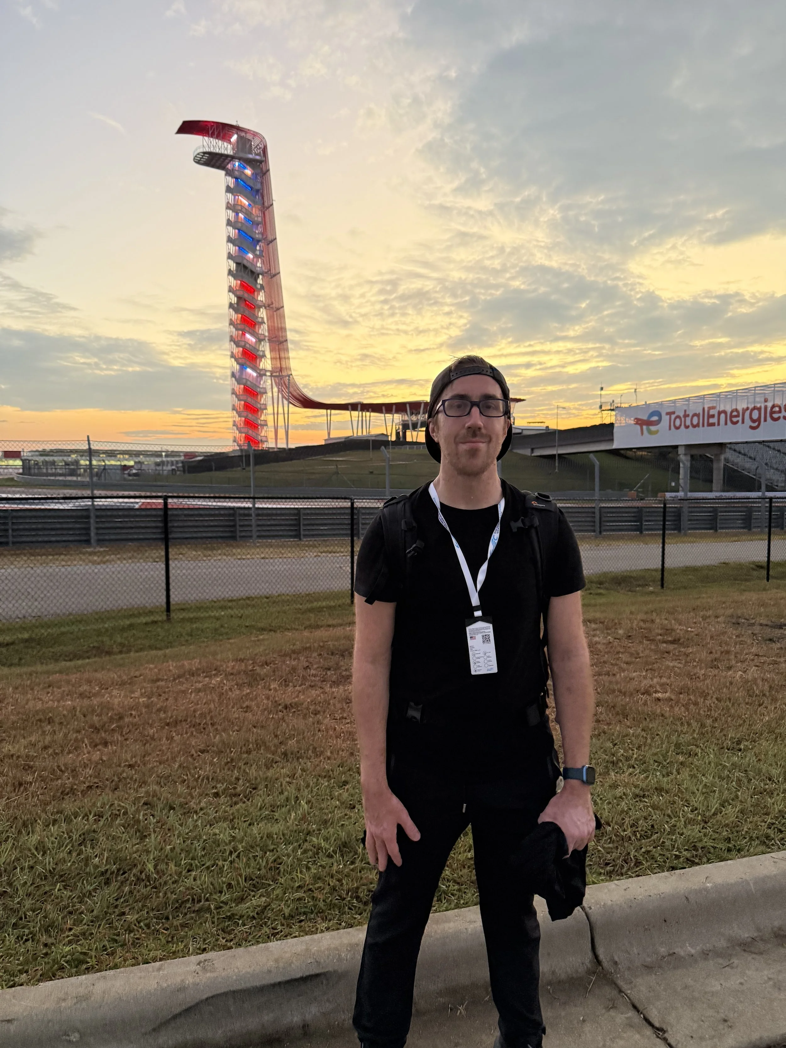 A man wearing black clothing and glasses standing on a sidewalk at a race track, with a tall, illuminated racing circuit tower and sunset sky in the background.