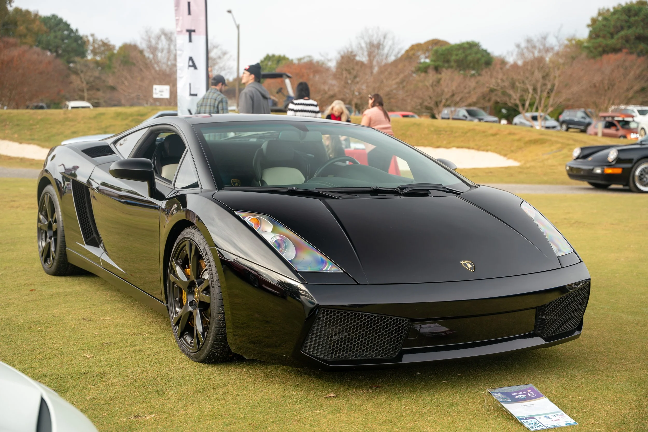 Black Lamborghini sports car displayed on a grassy field at a car show, with people and other vehicles in the background.