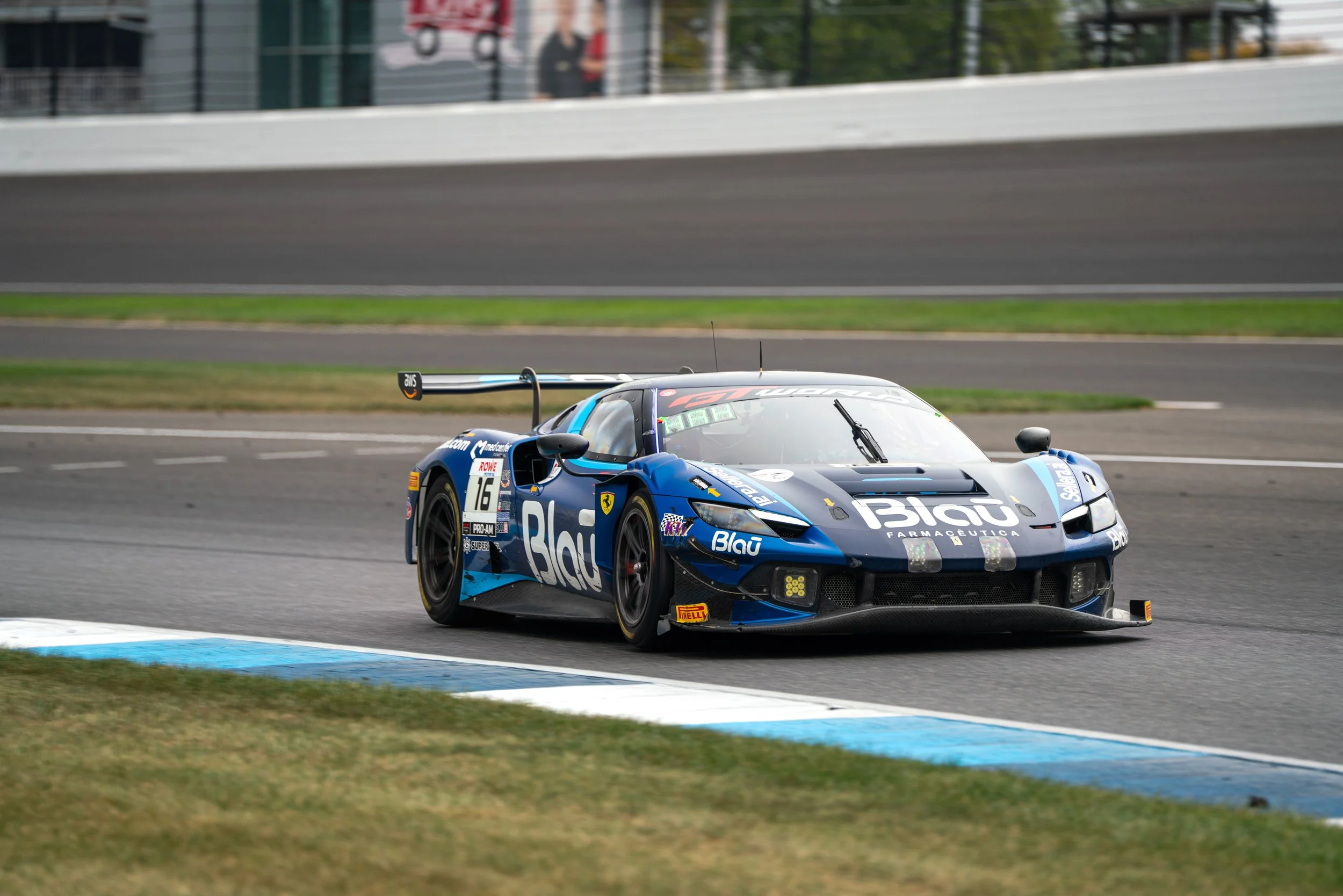 A blue and black race car, number 16, navigating a turn on a racing circuit. The car features sponsor logos, including 'Blau' and 'Pirelli', with a prominent rear wing and aerodynamic design.
