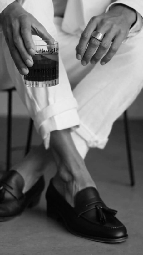 A flâneur sitting with crossed legs, wearing black dress shoes and white pants. They are holding a Negroni cocktail in one hand and resting the other hand on their knee.