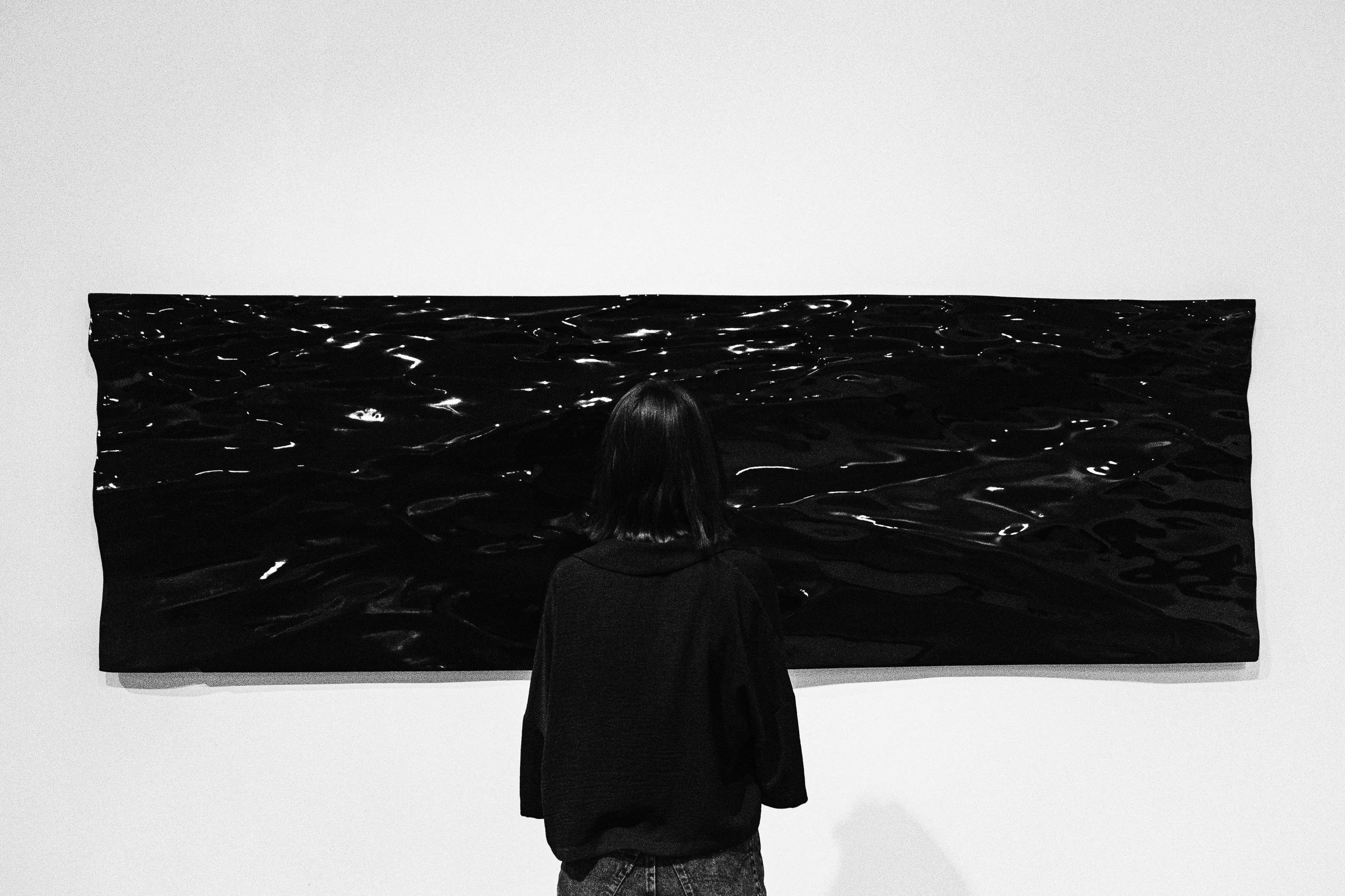 Person with shoulder-length hair view black abstract painting of water on gallery wall.