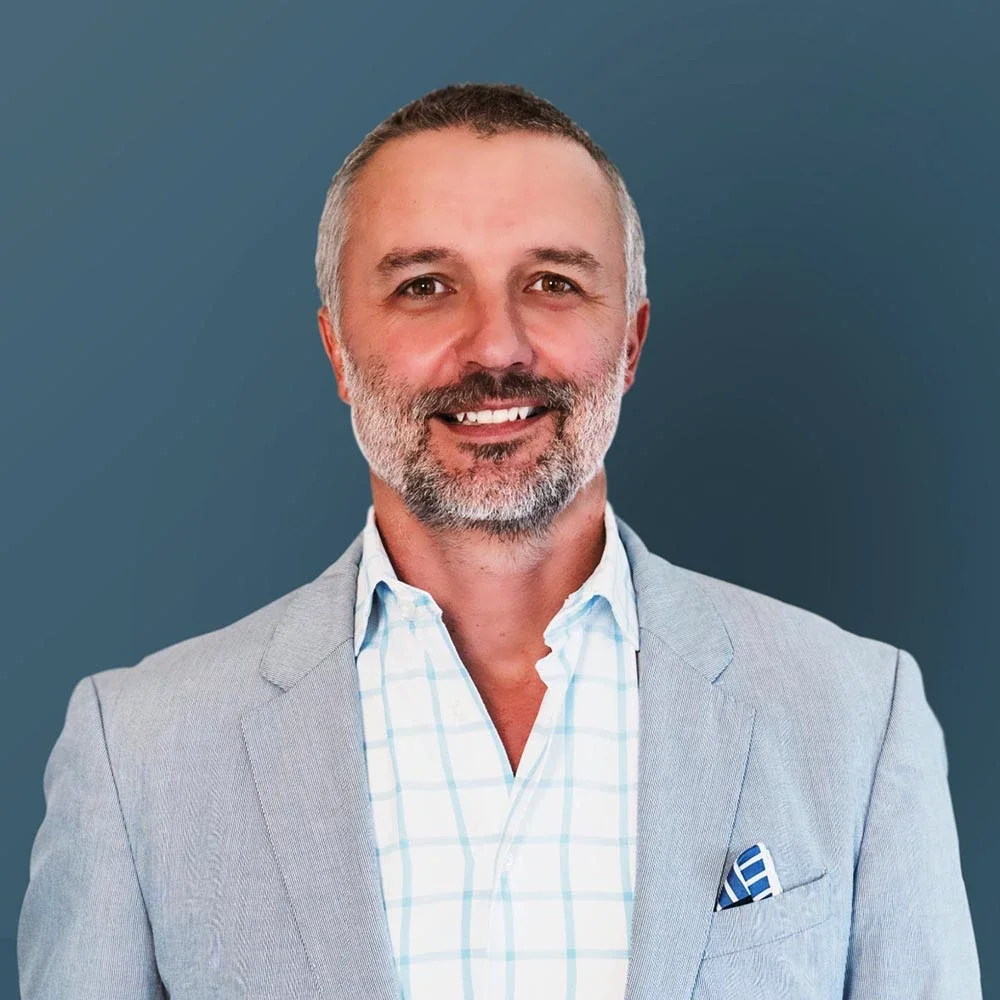 A smiling middle-aged man with a beard, wearing a light gray blazer over a white checkered shirt with a pocket square, against a plain blue background.