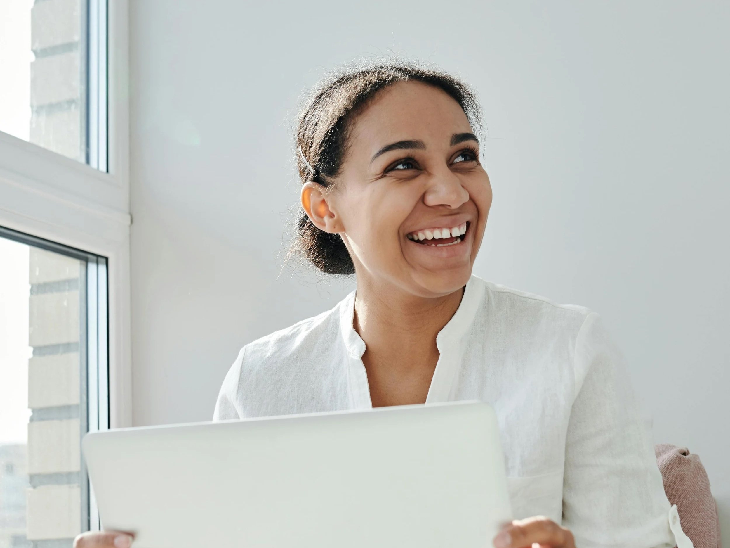 Young woman smiling and holding a tablet in a bright room near a window.