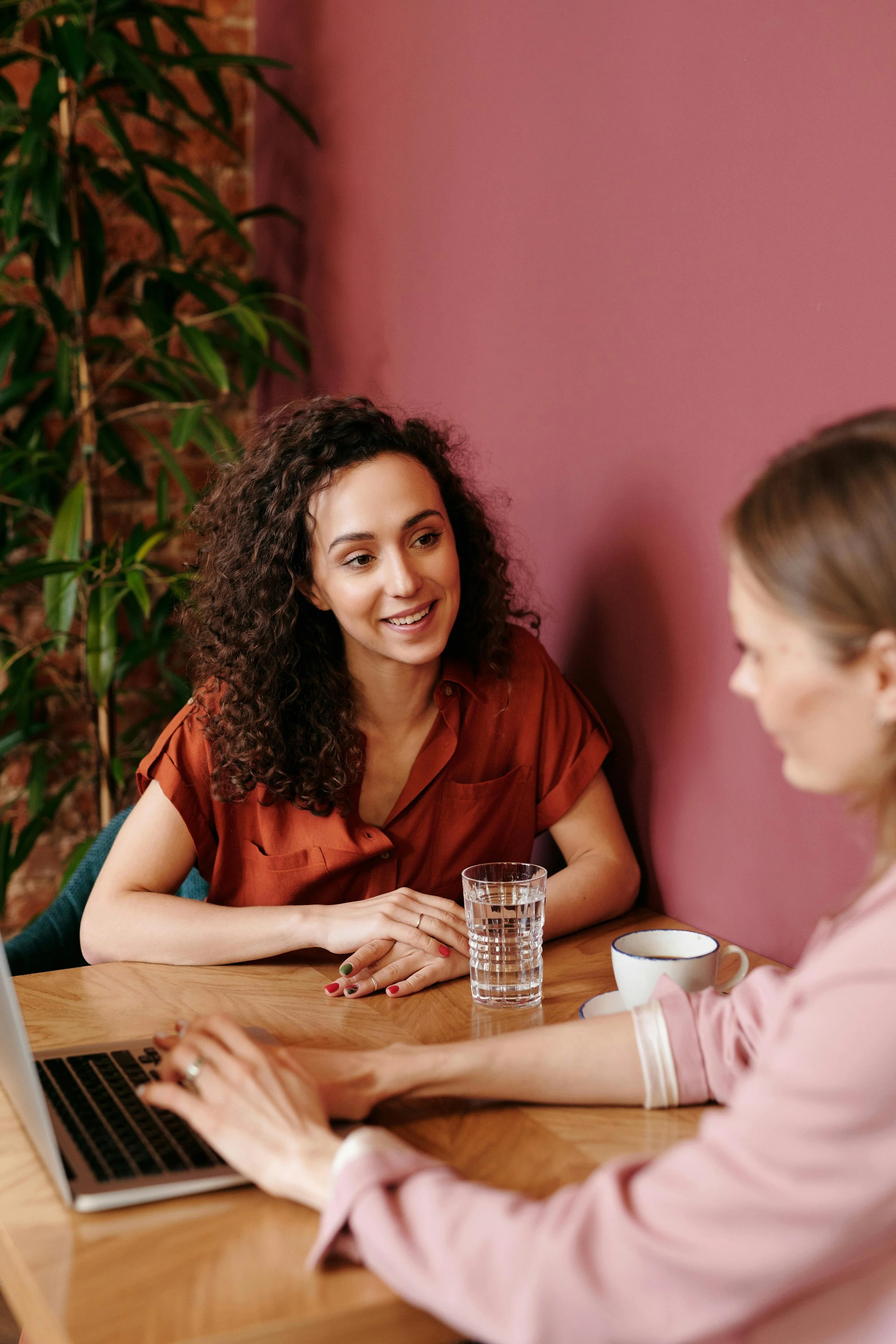 Two women having a conversation at a wooden table in a cozy room with a pink wall and a tall plant in the background. One woman with curly hair is smiling, and the other woman is working on a laptop.