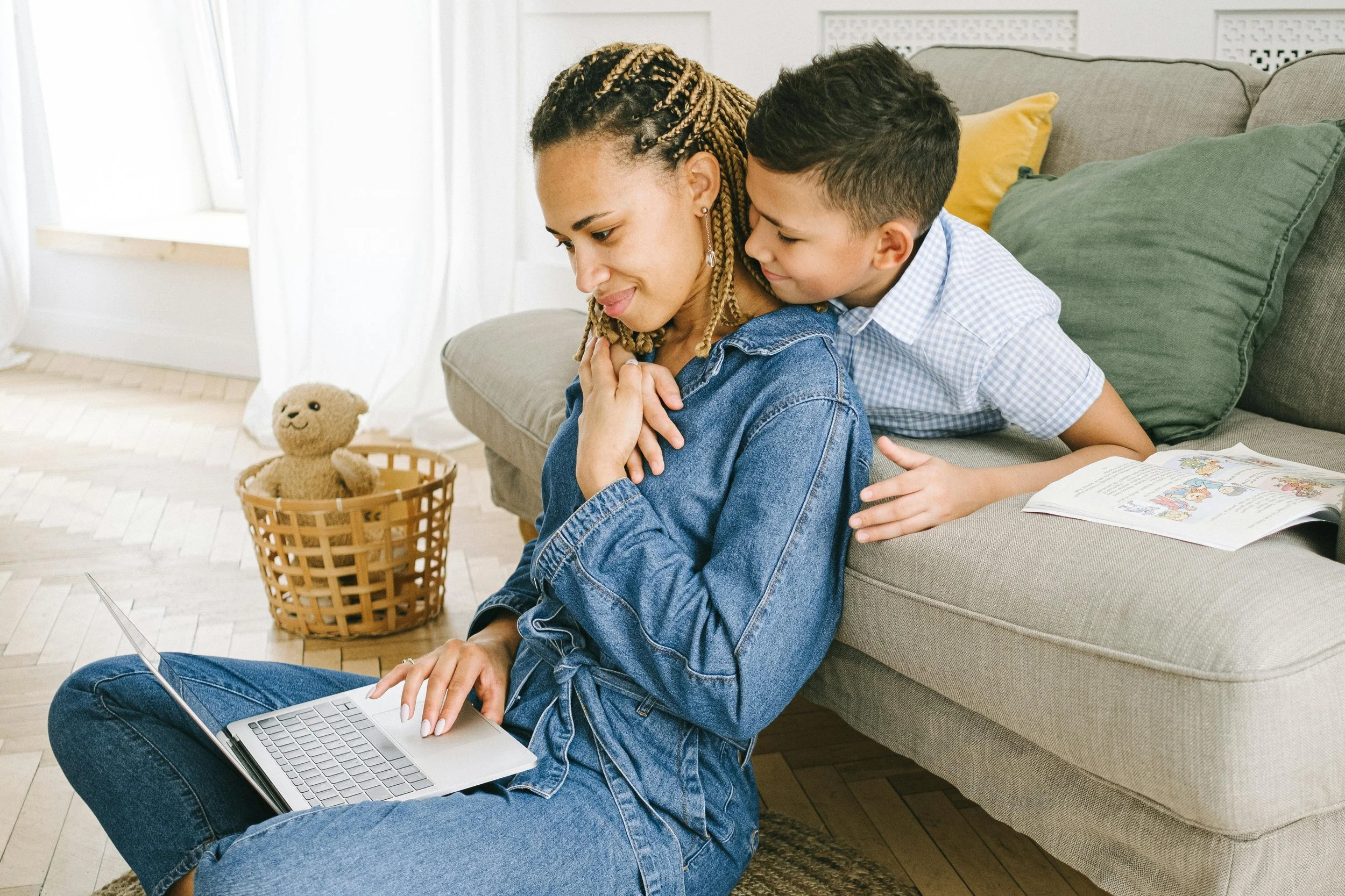 A woman with braided hair and a young boy on a living room sofa, with the boy leaning over the woman who is using a laptop on the floor. There is a basket with a teddy bear and a book open on the sofa.