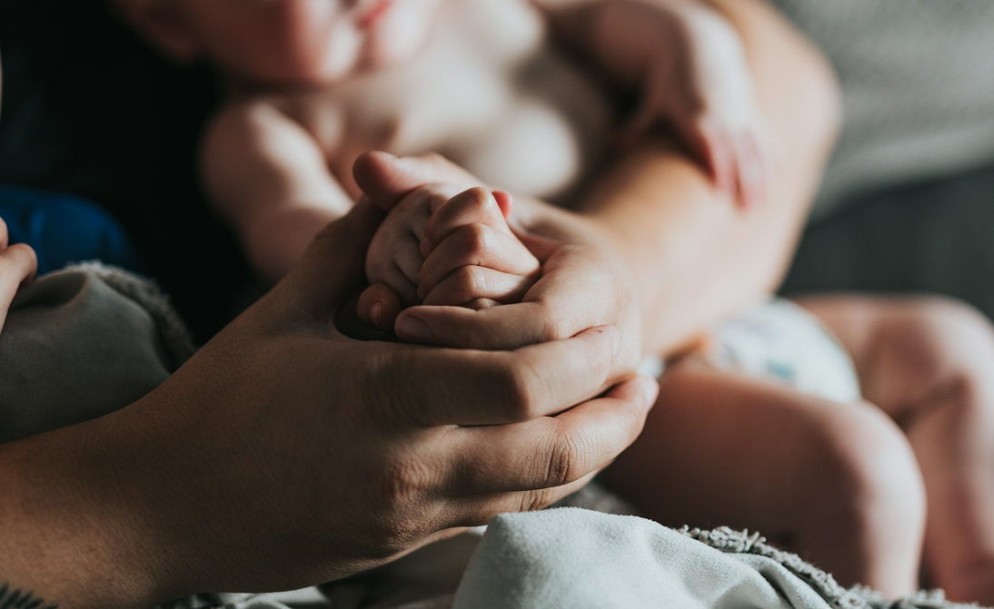 An adult holding a baby's hand, with the baby lying on a soft surface in the background.