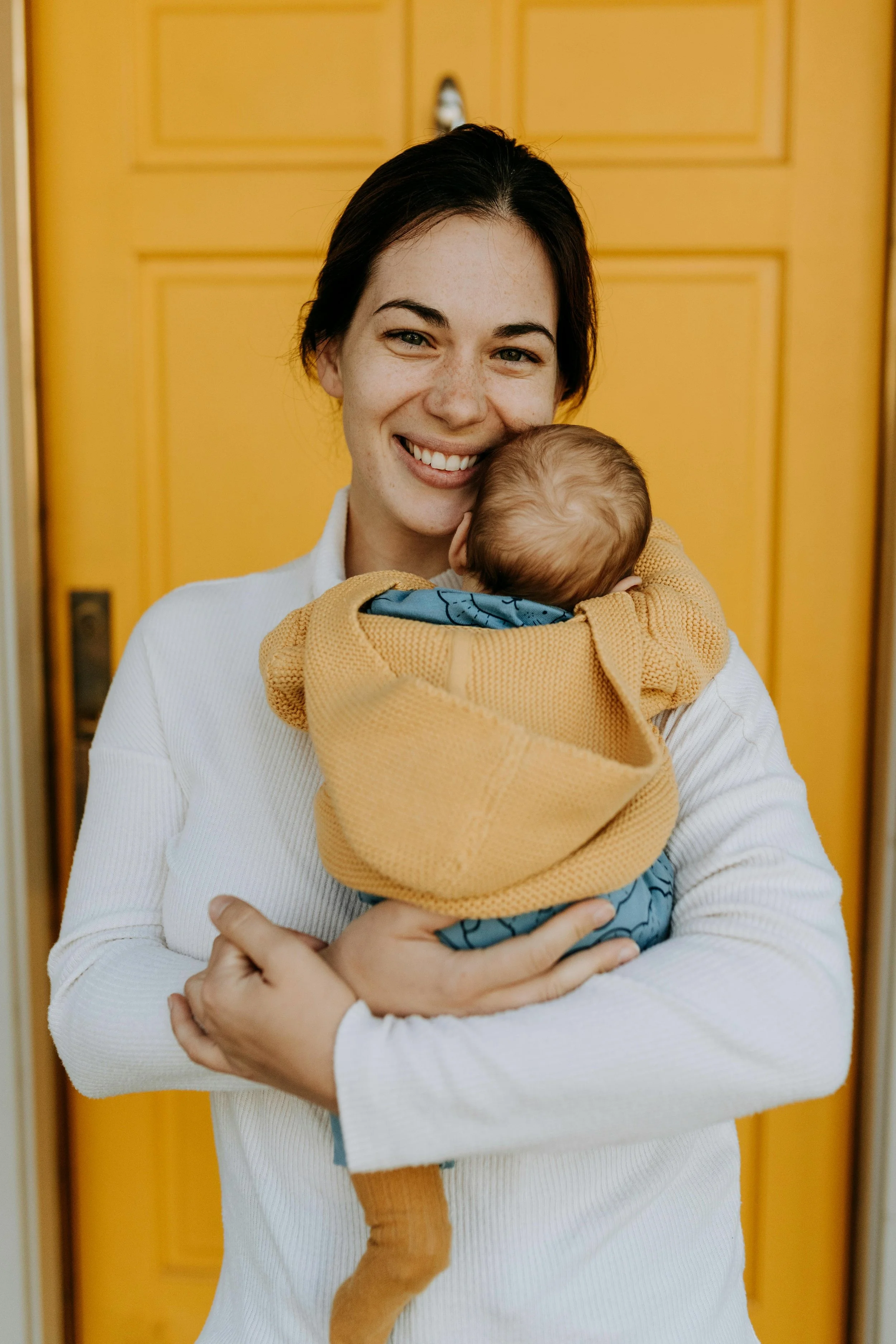 A woman smiling while holding a baby wrapped in a blanket in front of a yellow door.