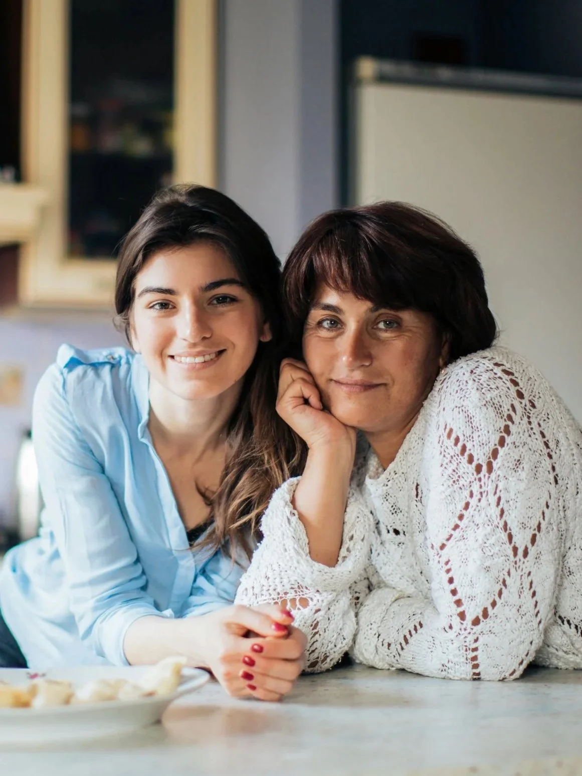 A young woman and an older woman are smiling and sitting at a table in a kitchen. The young woman has long dark hair and is wearing a light blue shirt, while the older woman has short dark hair and is wearing a white crochet sweater. They are close together with their hands clasped on the table.