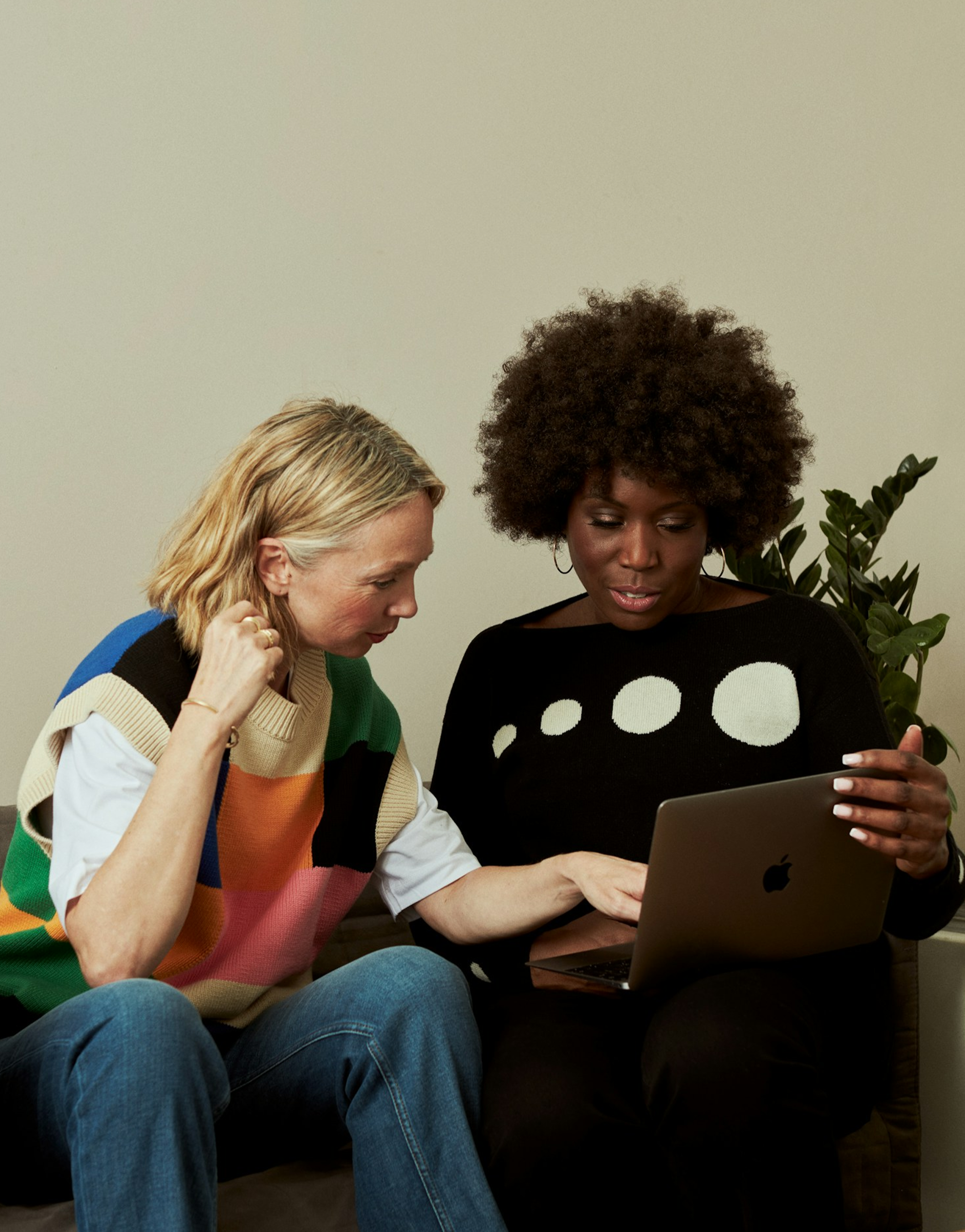 Two women sitting close together, looking at a laptop screen, one with blond hair in a colorful vest and the other with curly hair in a black sweater with white circles. They appear to be discussing something on the laptop.