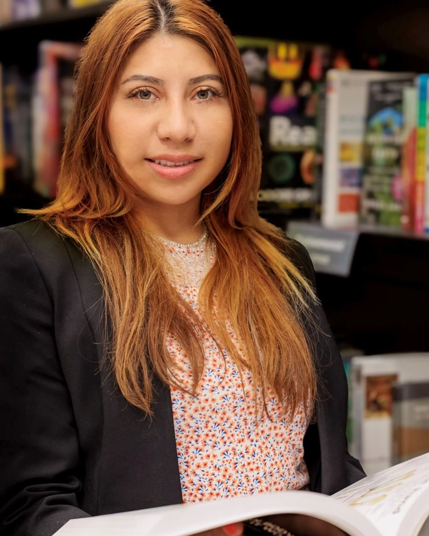Mujer con cabello largo y rubio, vestida con blusa de estampado floral y chaqueta negra, sosteniendo un libro abierto en un entorno de librería.
