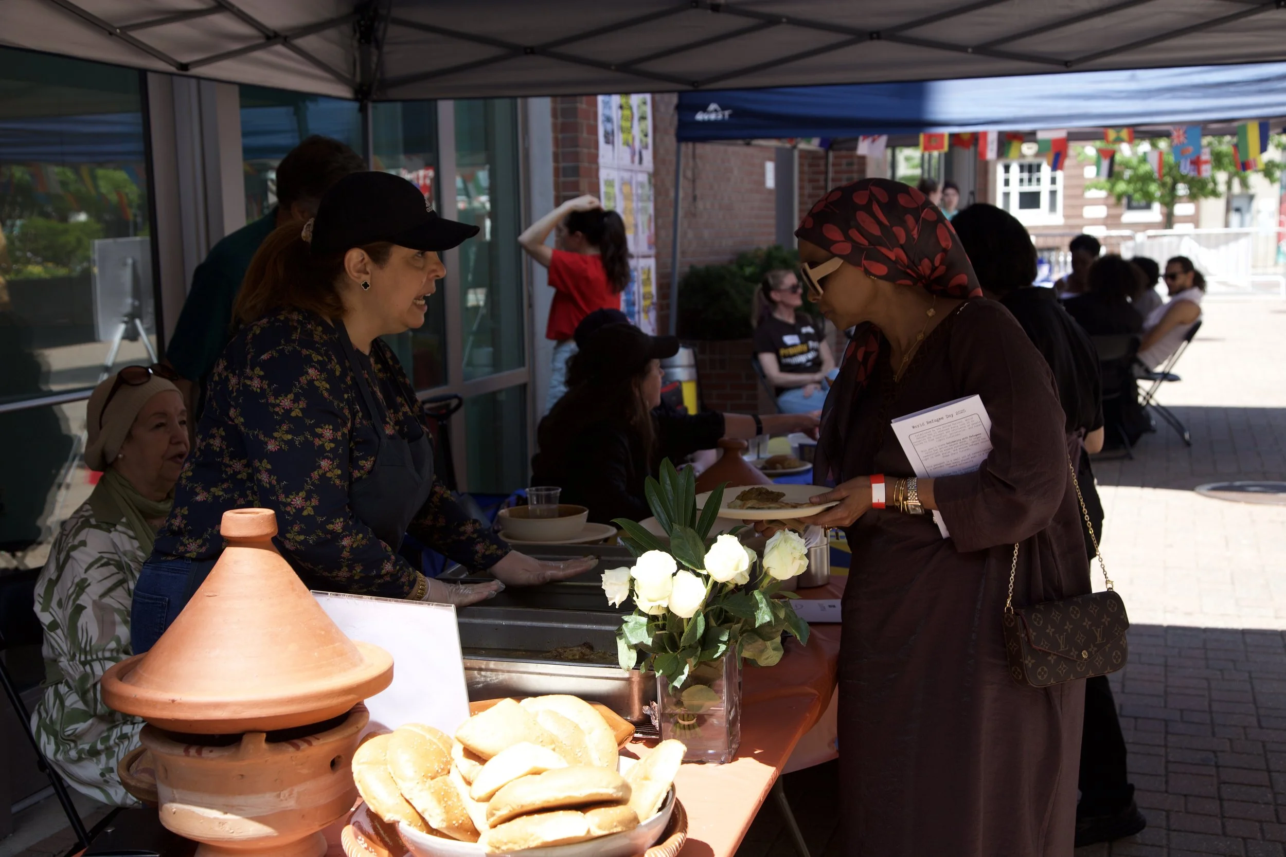 People at an outdoor event under a tent, with one woman serving food to another. There are flowers and bread on the table with flags hanging in the background.