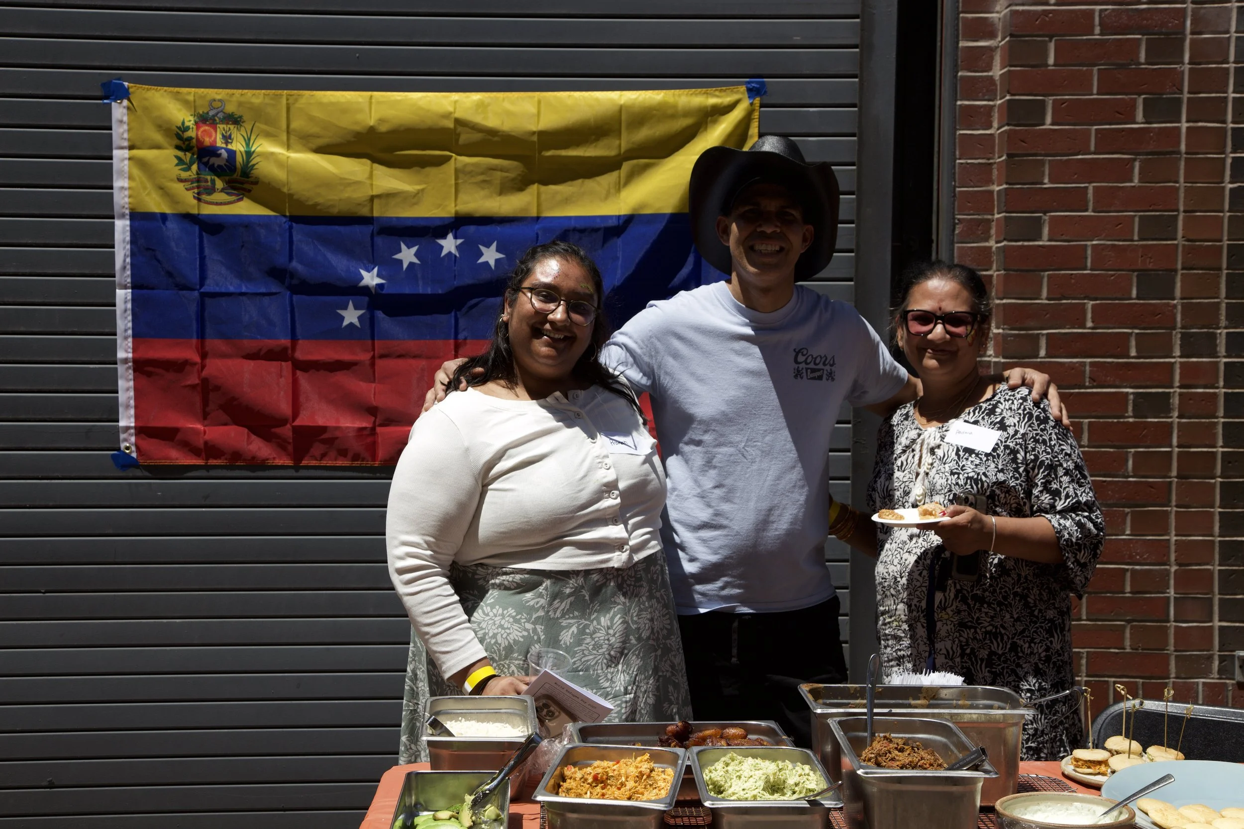 Three people standing together behind a table of food, with a Venezuelan flag in the background. The man in the middle is wearing a cowboy hat and white T-shirt, smiling. The woman on the left wears glasses and a light-colored top. The woman on the r