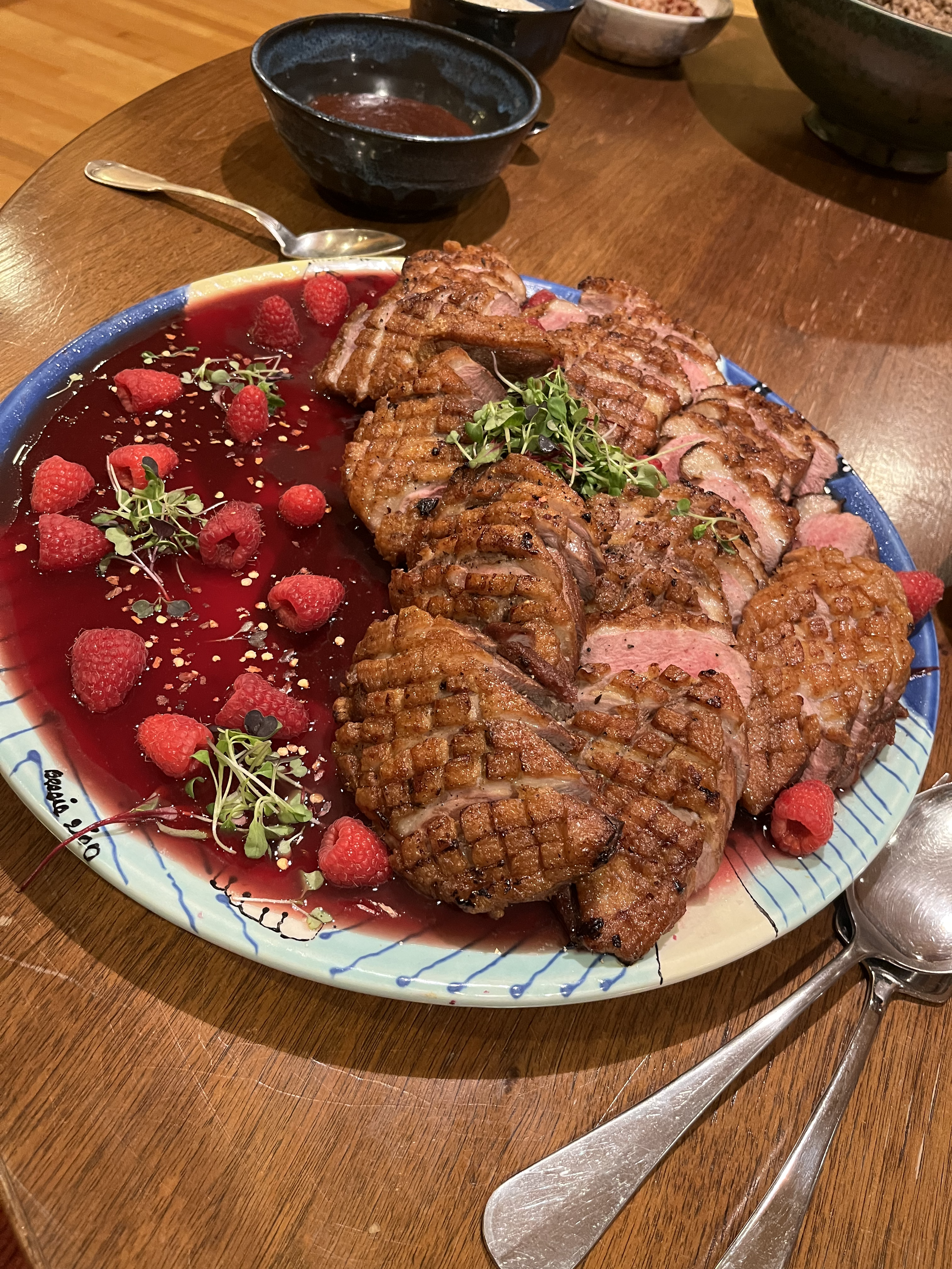 A large plate of assorted grilled meats served with raspberry sauce and garnished with raspberries and microgreens, on a wooden table.