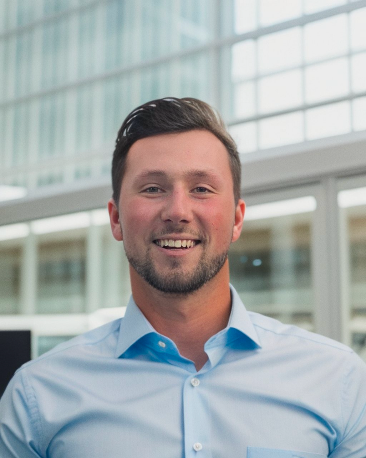 Smiling man in a light blue shirt with a modern office building background.