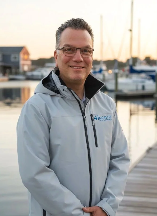 Man wearing glasses and a light gray jacket standing on a dock at sunset with boats in the background.