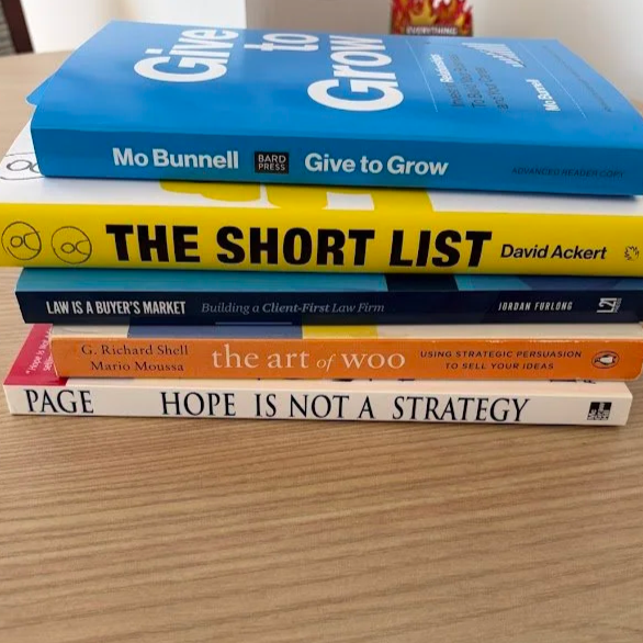 Stack of five books on a wooden surface, with a blurred background including a can and a container. The books are titled 'Give to Grow' by Mo Bunnell, 'The Short List' by David Ackert, 'Law is a Buyer's Market' by Jordan Furlong, 'The Art of Woo' by G. Richard Shell and Mario Moussa, and 'Hope is Not a Strategy' by PAGE.