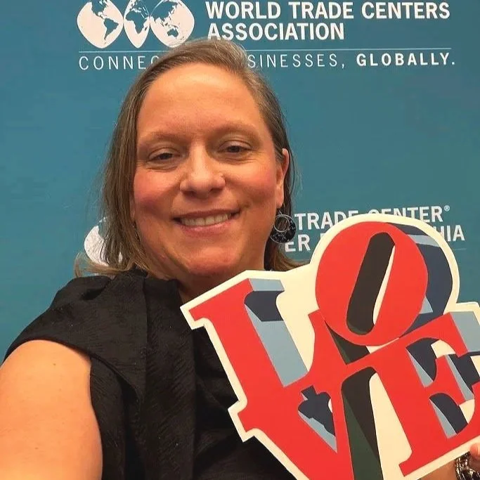 Woman smiling at a conference holding a colorful 'LOVE' sign with a blue background that includes the words 'World Trade Centers Association' and 'Connecting Businesses, Globally.'