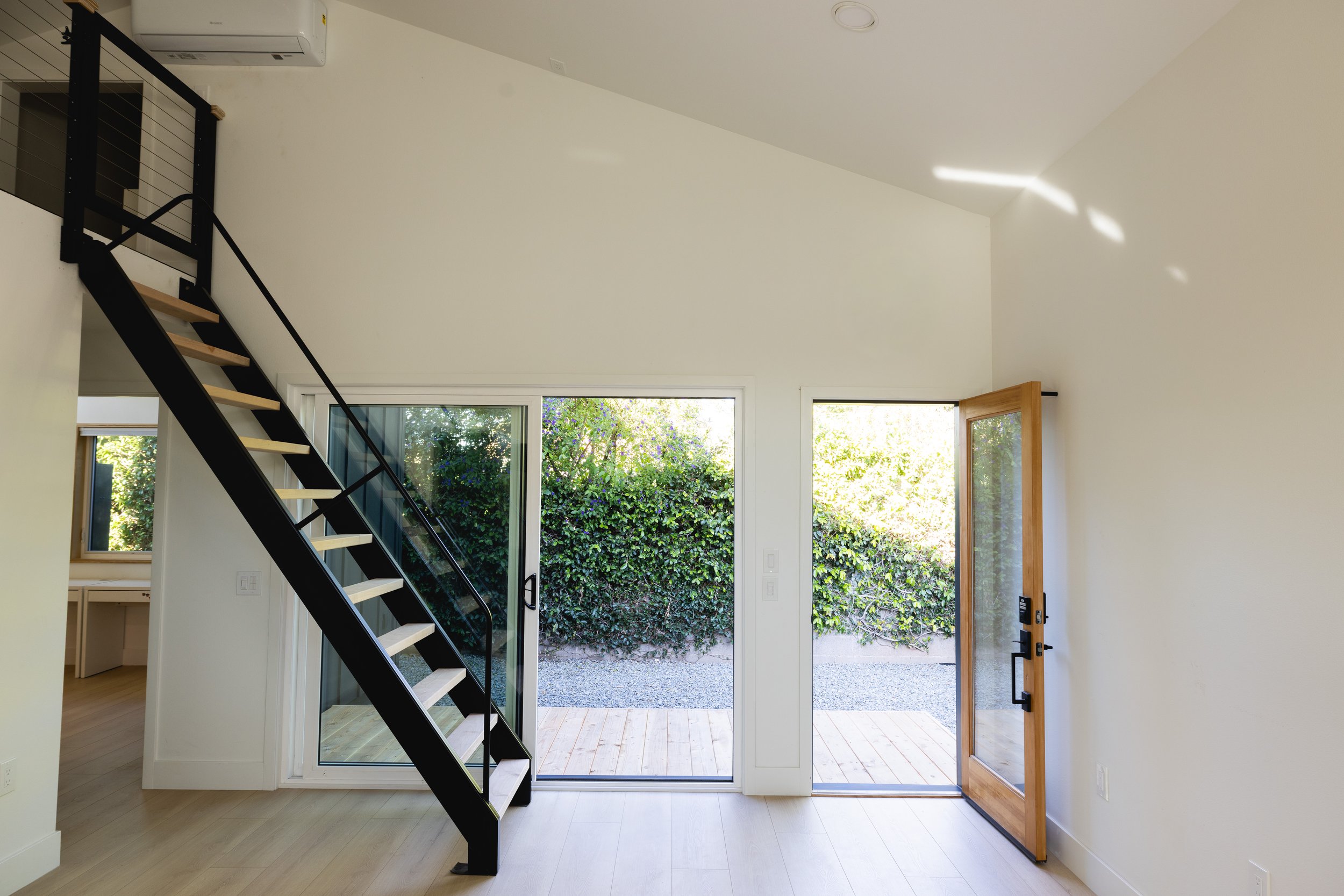 From inside the ADU looking out to the yard where a large vine plant encompasses a fence. Inside the ADU a black steel staircase hangs down from the loft space into the kitchen/living area
