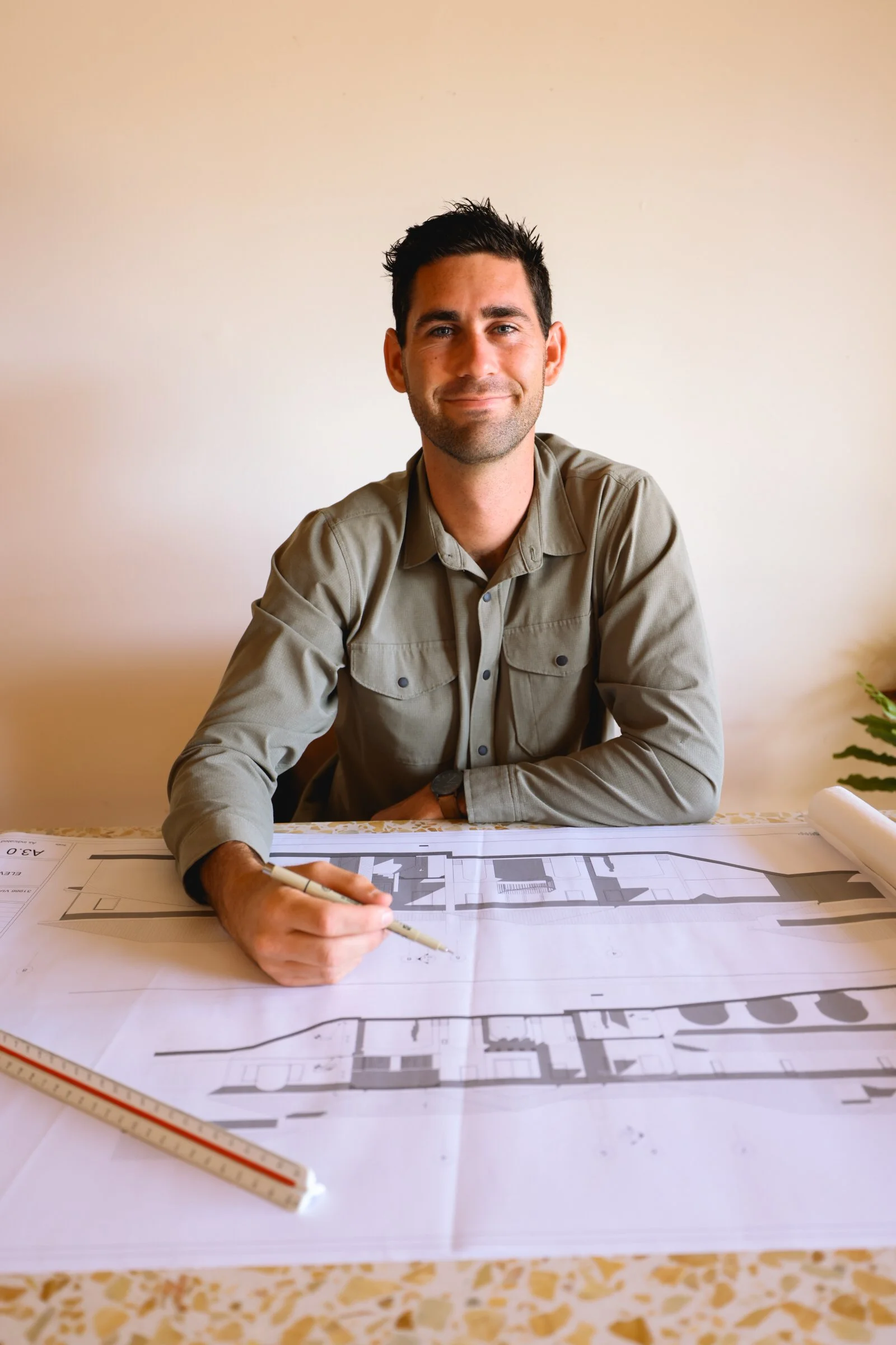 Man sitting at a table with architectural blueprints, holding a pen, smiling.