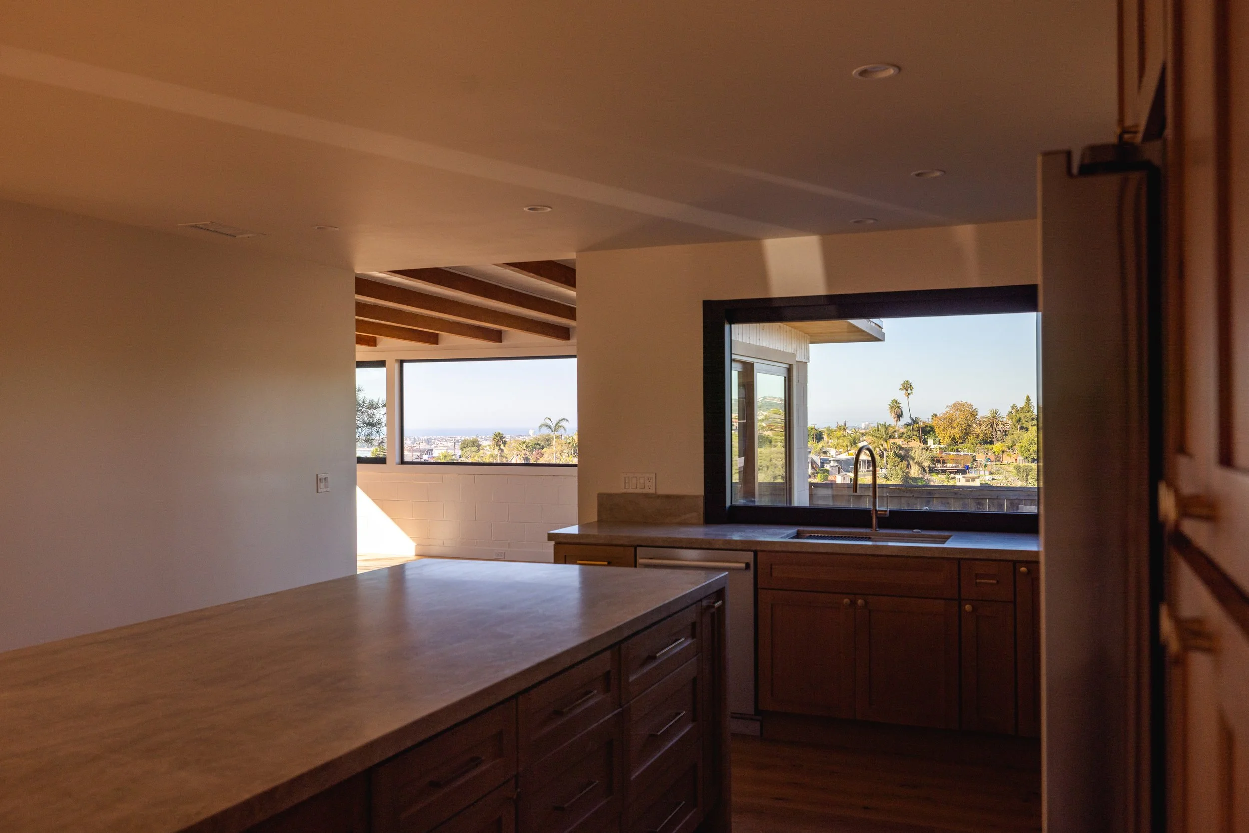 Image of a newly remodeled, modern, kitchen with paneled wood and a light marble countertop. Views of the San Diego Bay and palm trees are seen from the large window that opens and closes from the kitchen sink