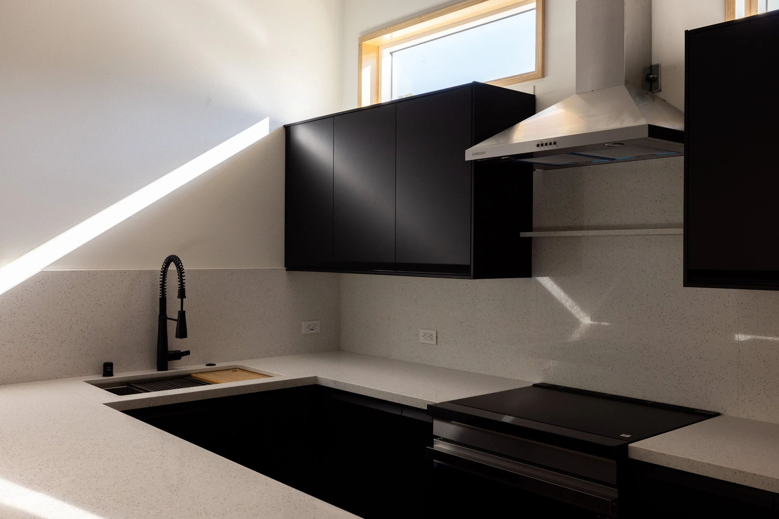 image of a modern kitchen, with black cabinets and sink, and crisp white countertops and paint, and a rectangular window above brining fresh bright light inside the ADU