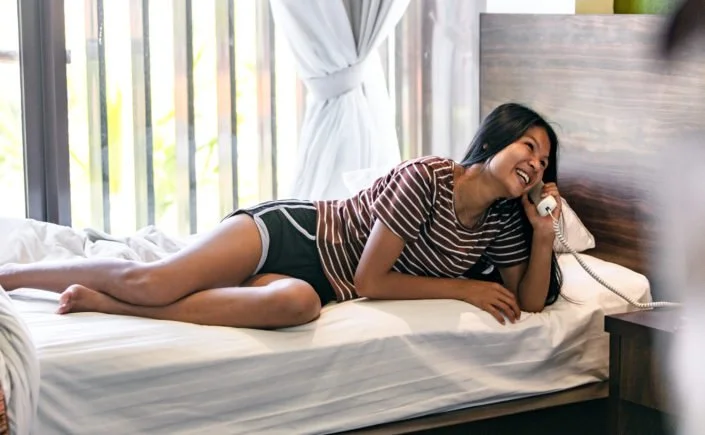 Woman lying on her side on a bed, talking on a landline phone, smiling, with a window and curtains in the background.
